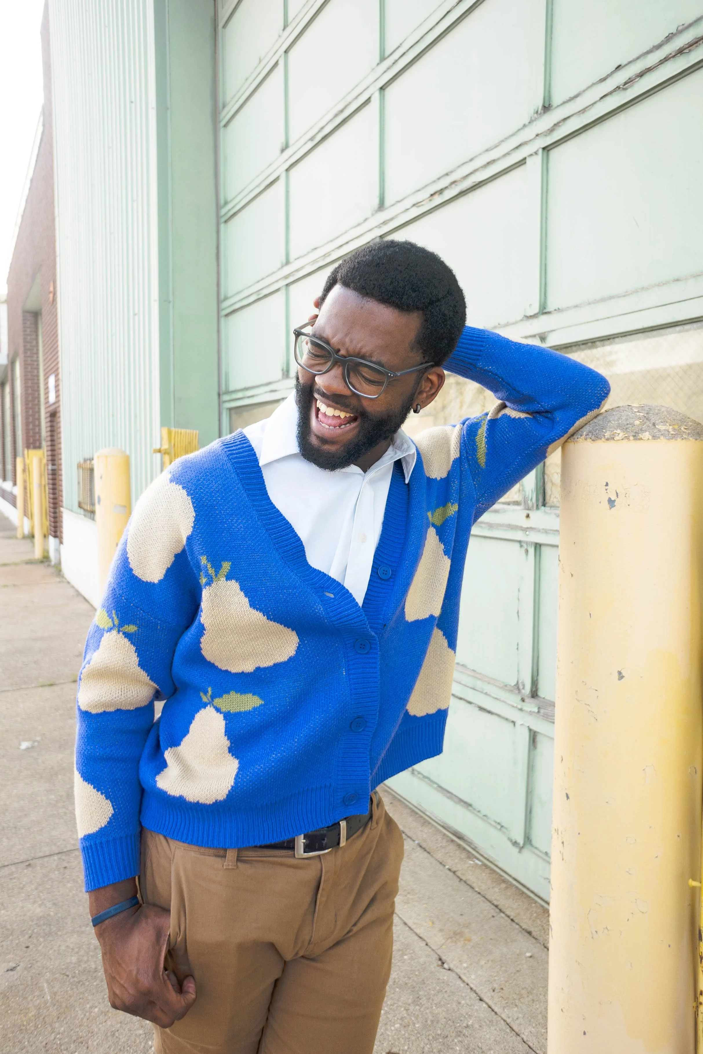 A man with glasses, a beard, and short hair, wearing a white shirt and a blue cardigan with large cream-colored pears on it, standing outdoors against a light green metal building, smiling and leaning on a yellow post.
