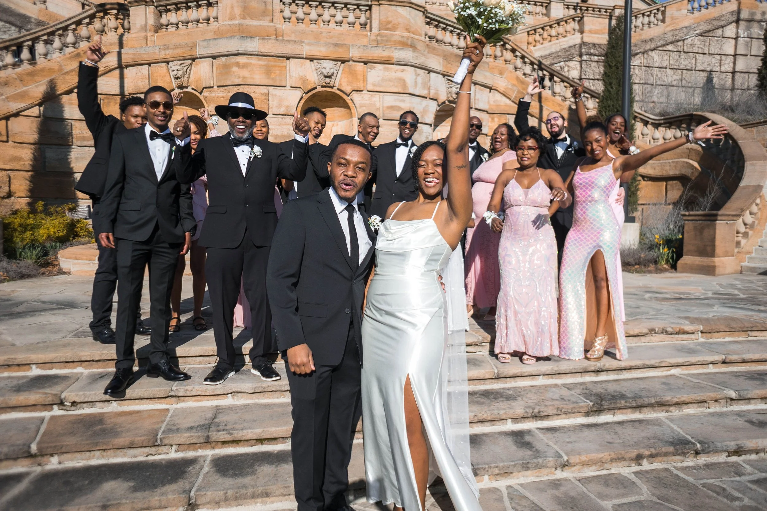 Wedding party with a bride in a white gown and a groom in a black suit, surrounded by bridesmaids in pink dresses and groomsmen in black tuxedos, standing on outdoor stone steps with a stone building in the background.