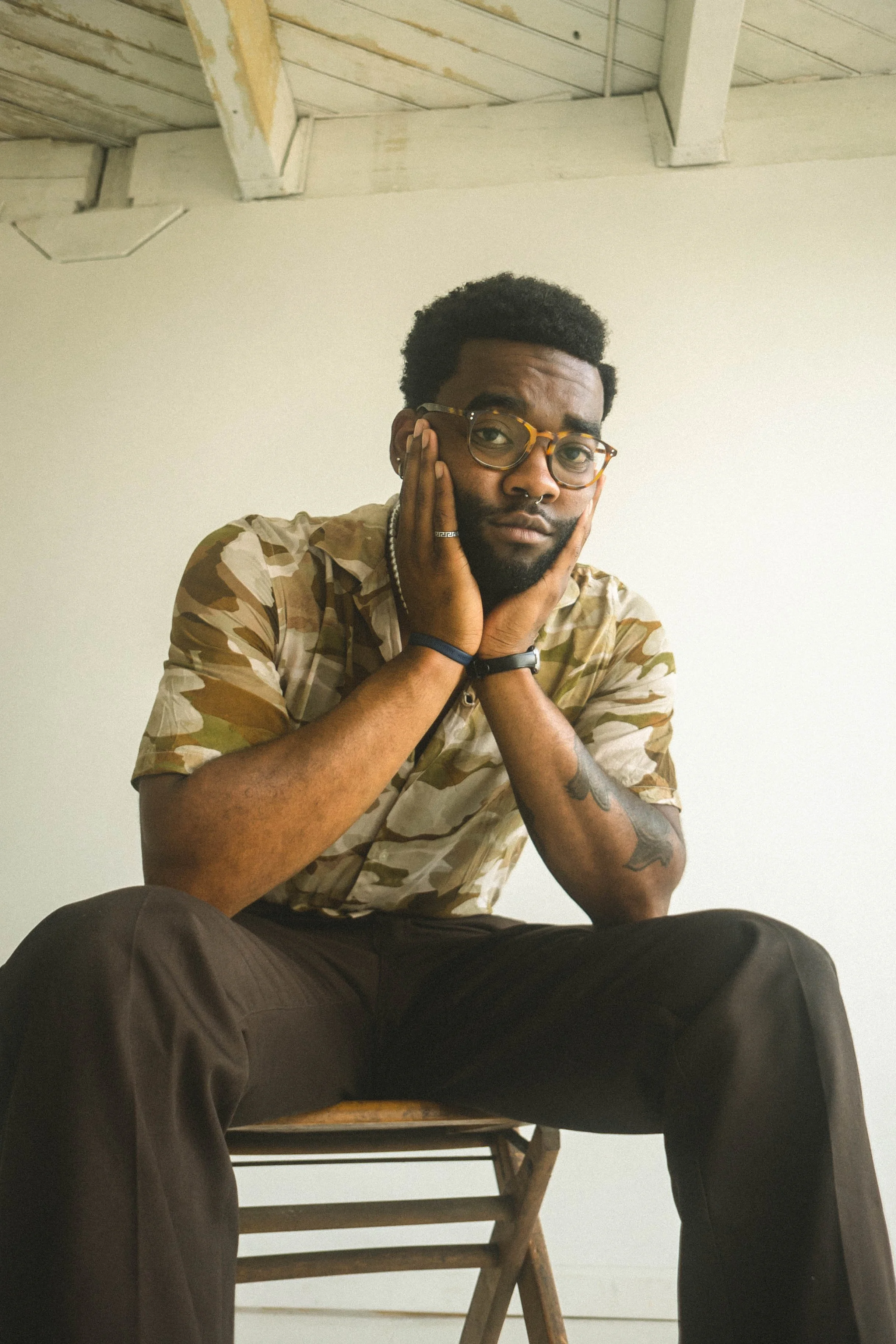 A man sitting on a wooden chair with his hands on his face, wearing glasses, a camouflage shirt, and dark pants, in a room with a white wall and wooden ceiling.