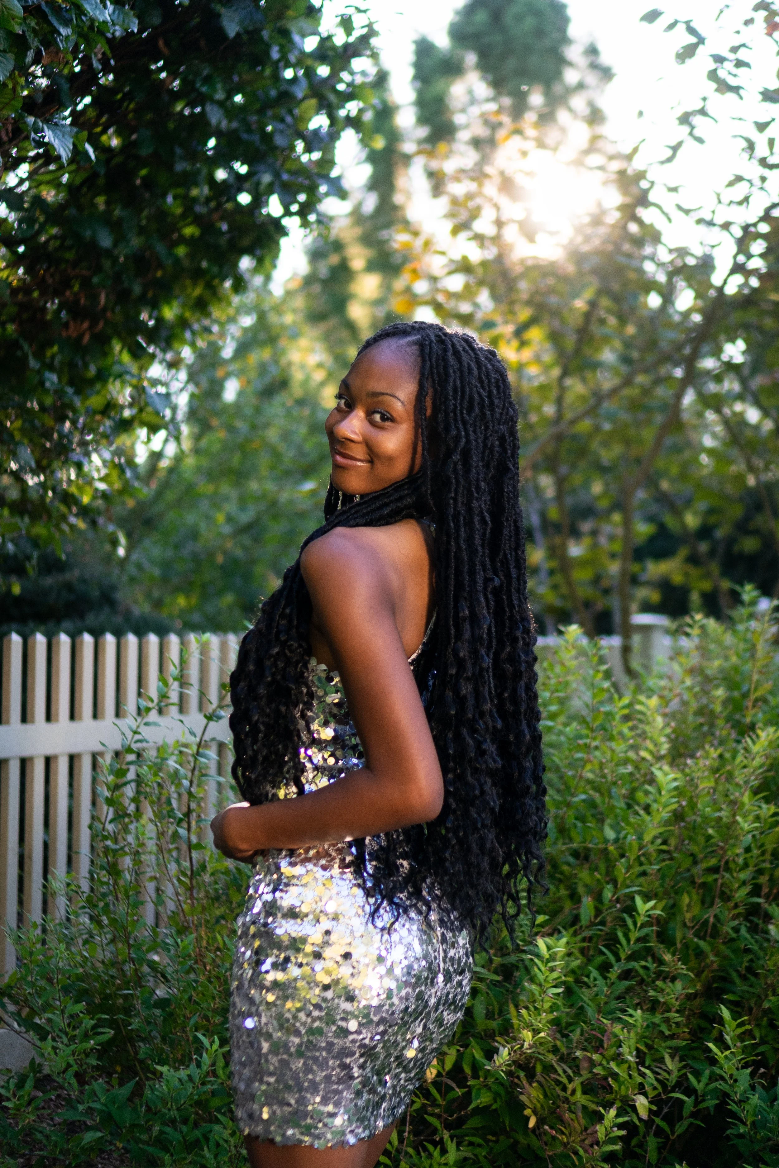 A woman with long, curly black hair wearing a sparkly silver dress stands outdoors in a garden with trees and greenery, smiling and looking over her shoulder during sunset.