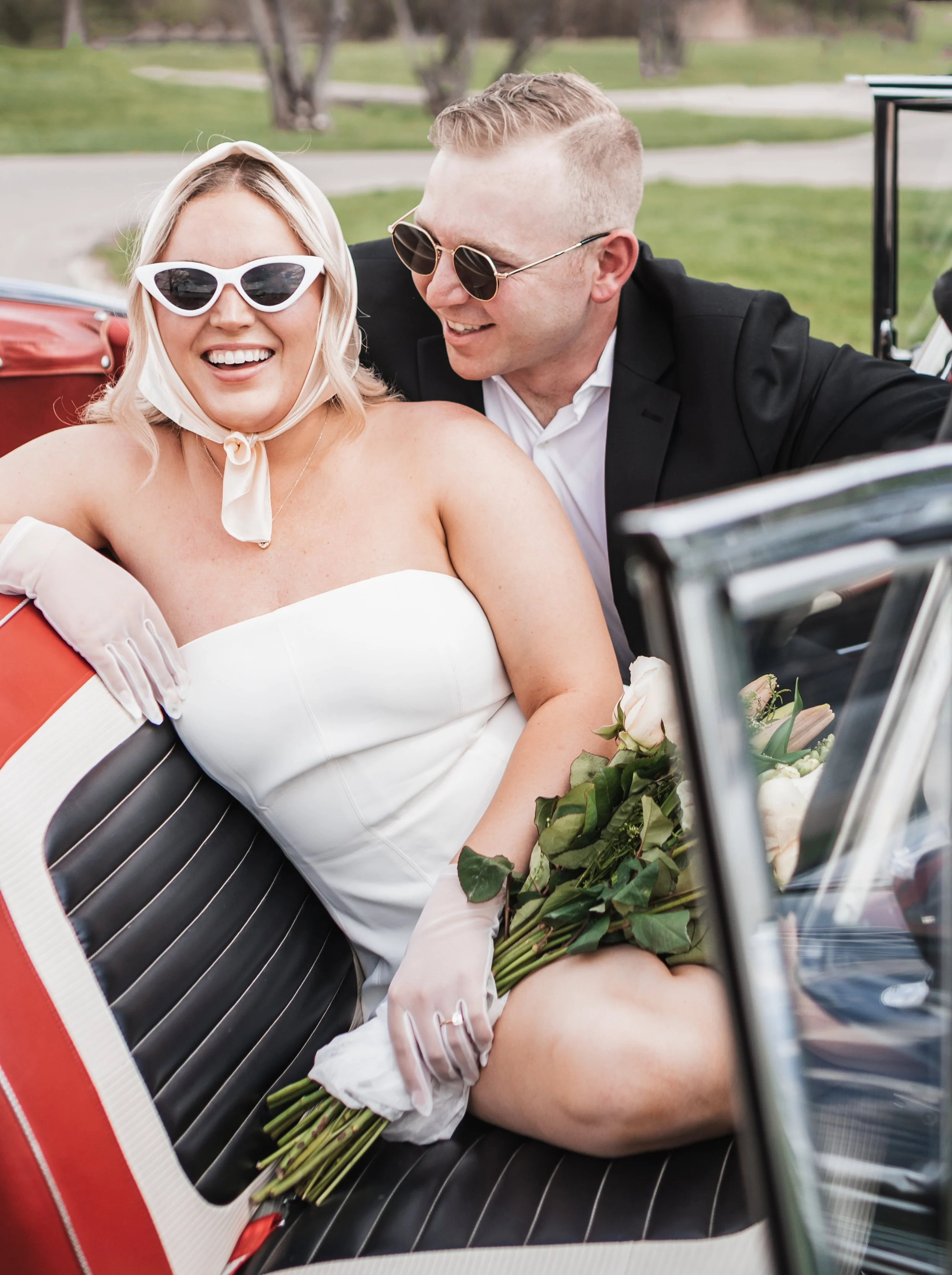 A woman in a wedding dress and sunglasses sitting in a vintage car, holding a bouquet of flowers, with a man in a black suit and sunglasses leaning towards her, both smiling and enjoying the moment outside on a grassy area.