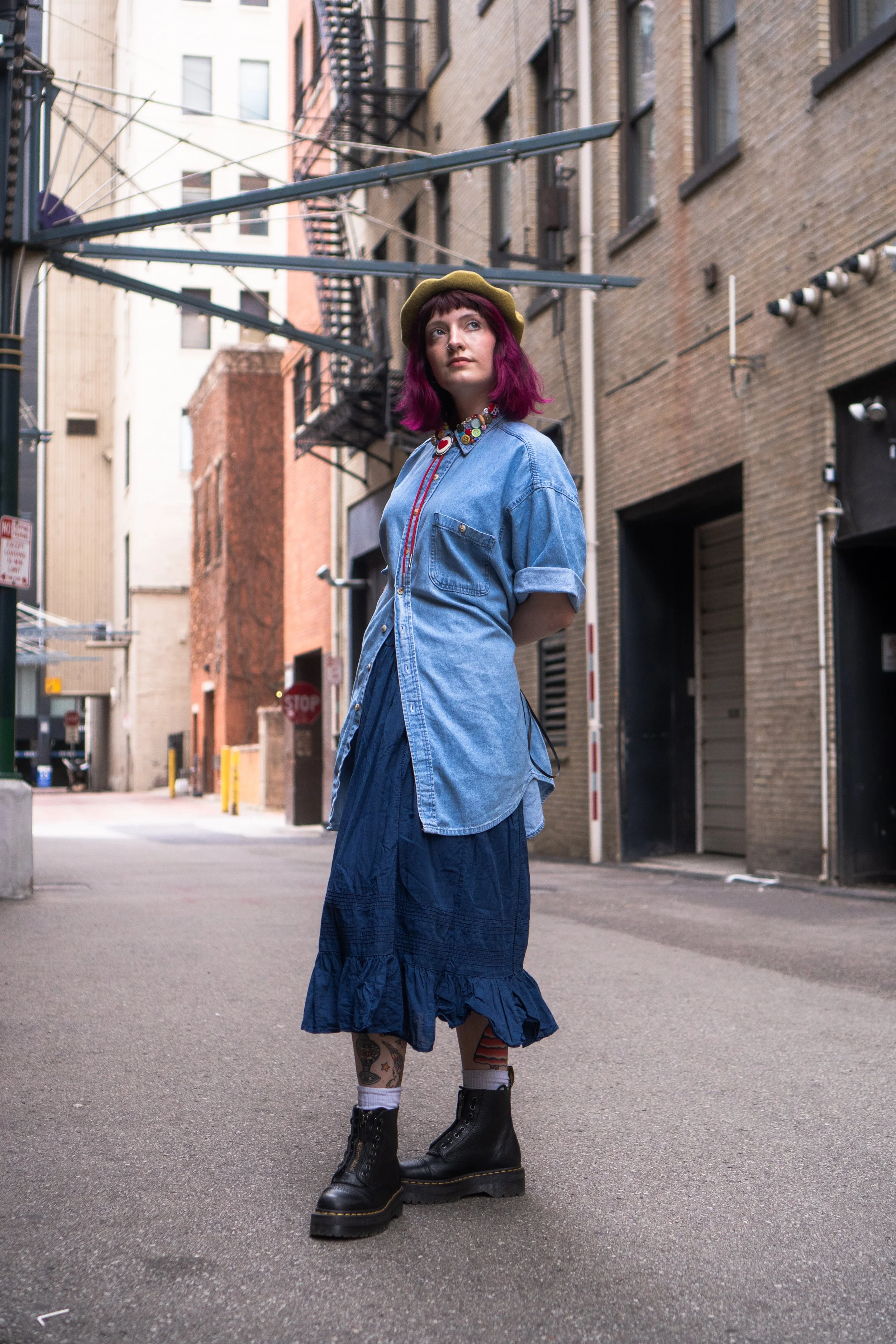 A woman with magenta hair wearing a green beret, denim shirt, and a blue skirt standing in an alleyway with brick and concrete buildings on either side.