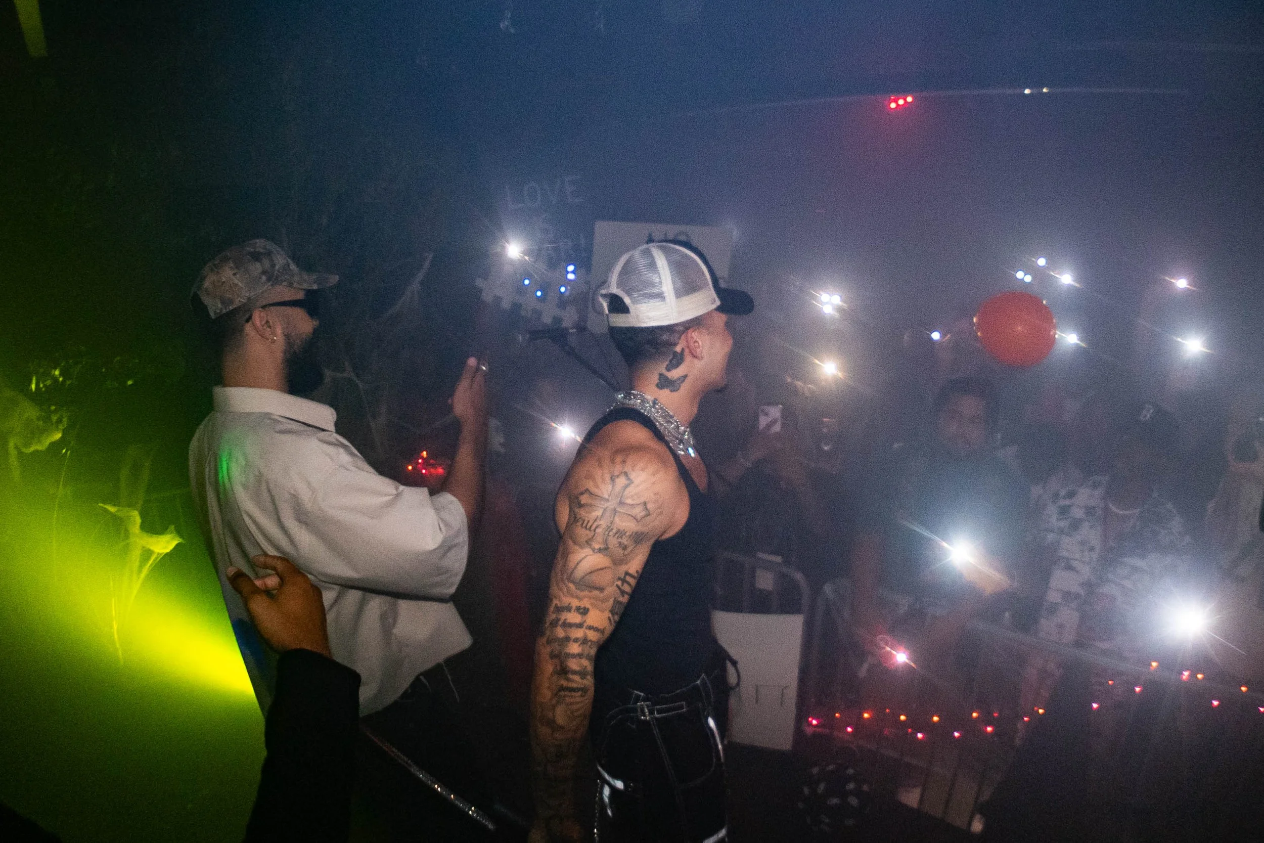 A person with tattoos and a cap standing at a crowded, dimly lit party with colorful lights and a red balloon.