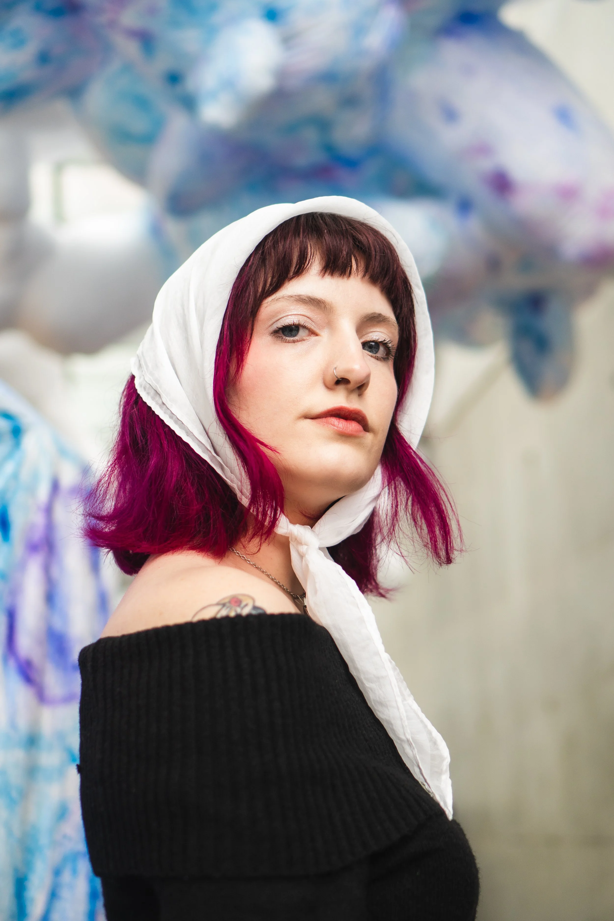 A young woman with shoulder-length dark red hair with purple tips, wearing a white headscarf, black off-shoulder sweater, and a necklace, standing in front of colorful balloons.
