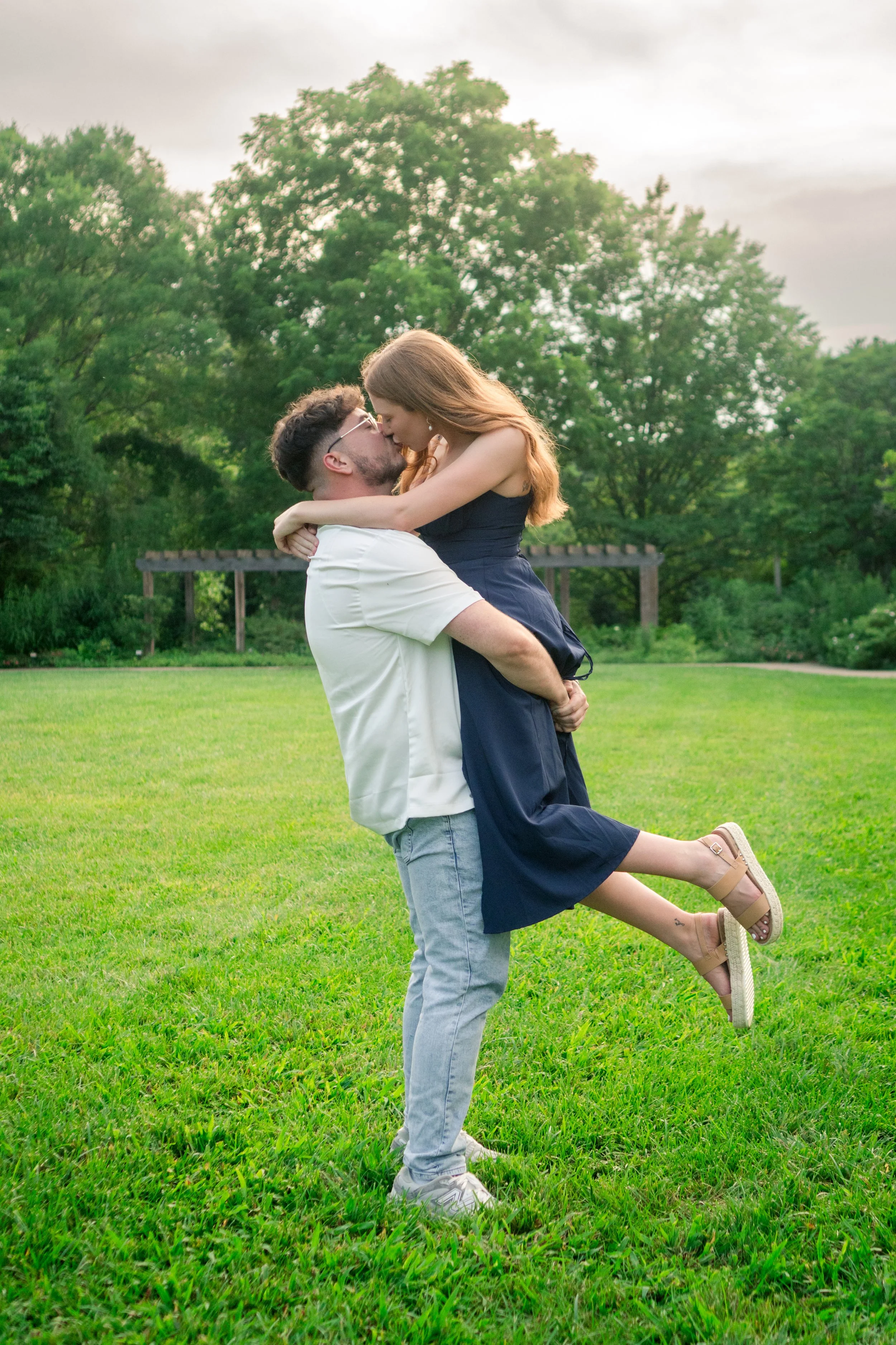 A man lifting a woman in a grassy park, both leaning in for a kiss, with trees and a cloudy sky in the background.