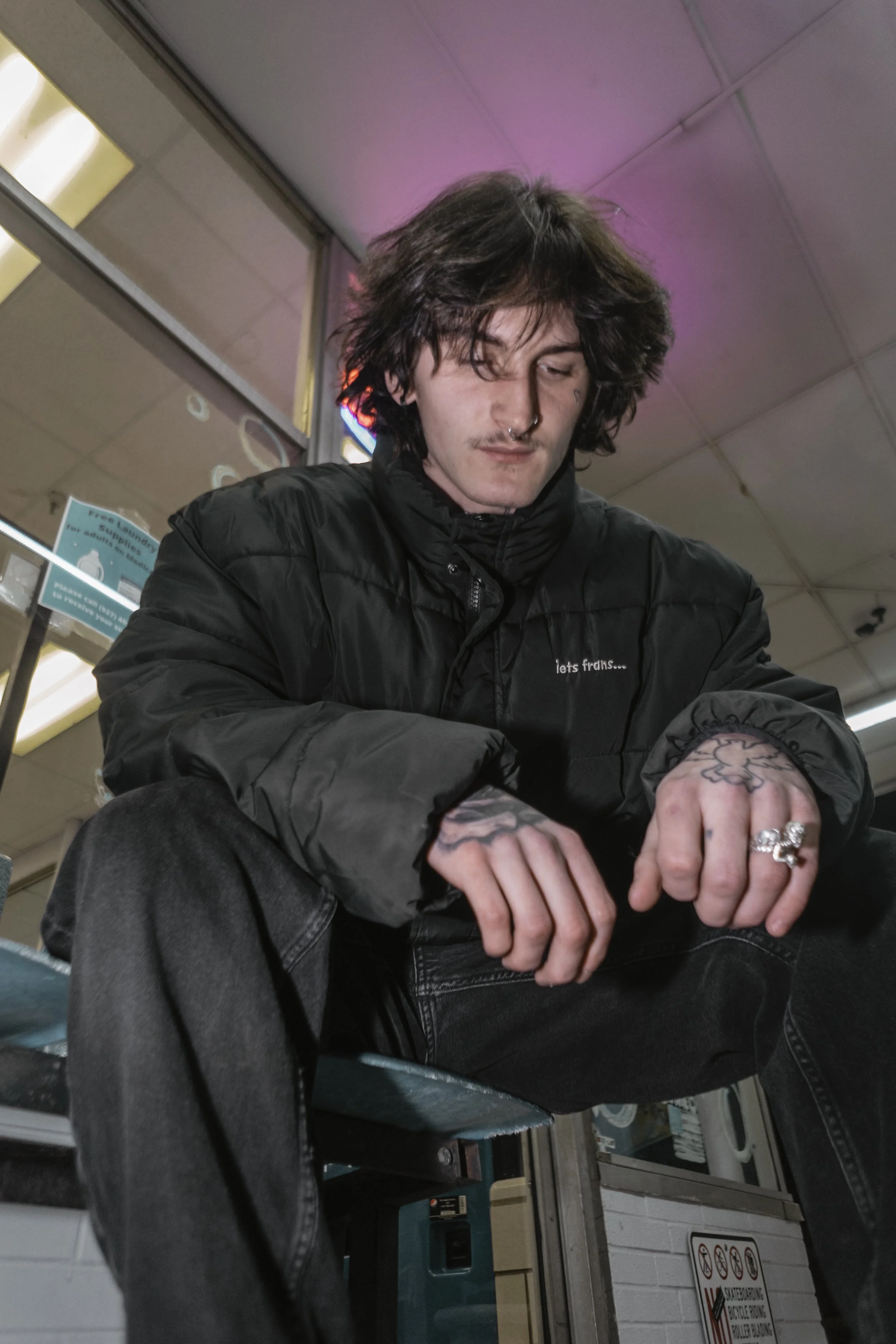 A young man with dark, messy hair and tattoos on his hands and fingers, sitting on a bench inside a store or waiting area, wearing a black jacket and dark jeans.