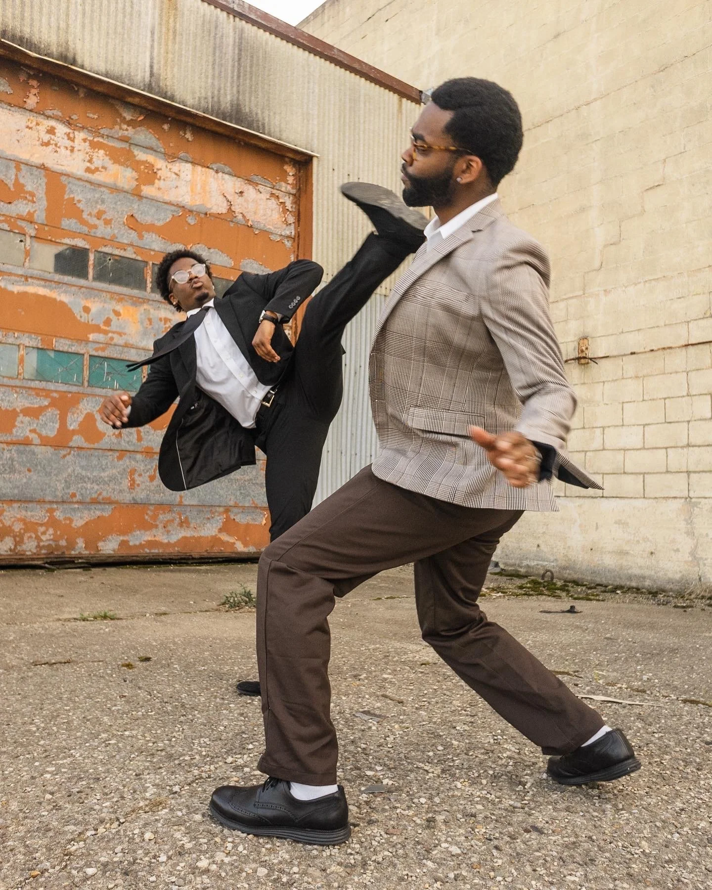 Two men in business suits fighting in an alleyway, one executing a high kick at the other.
