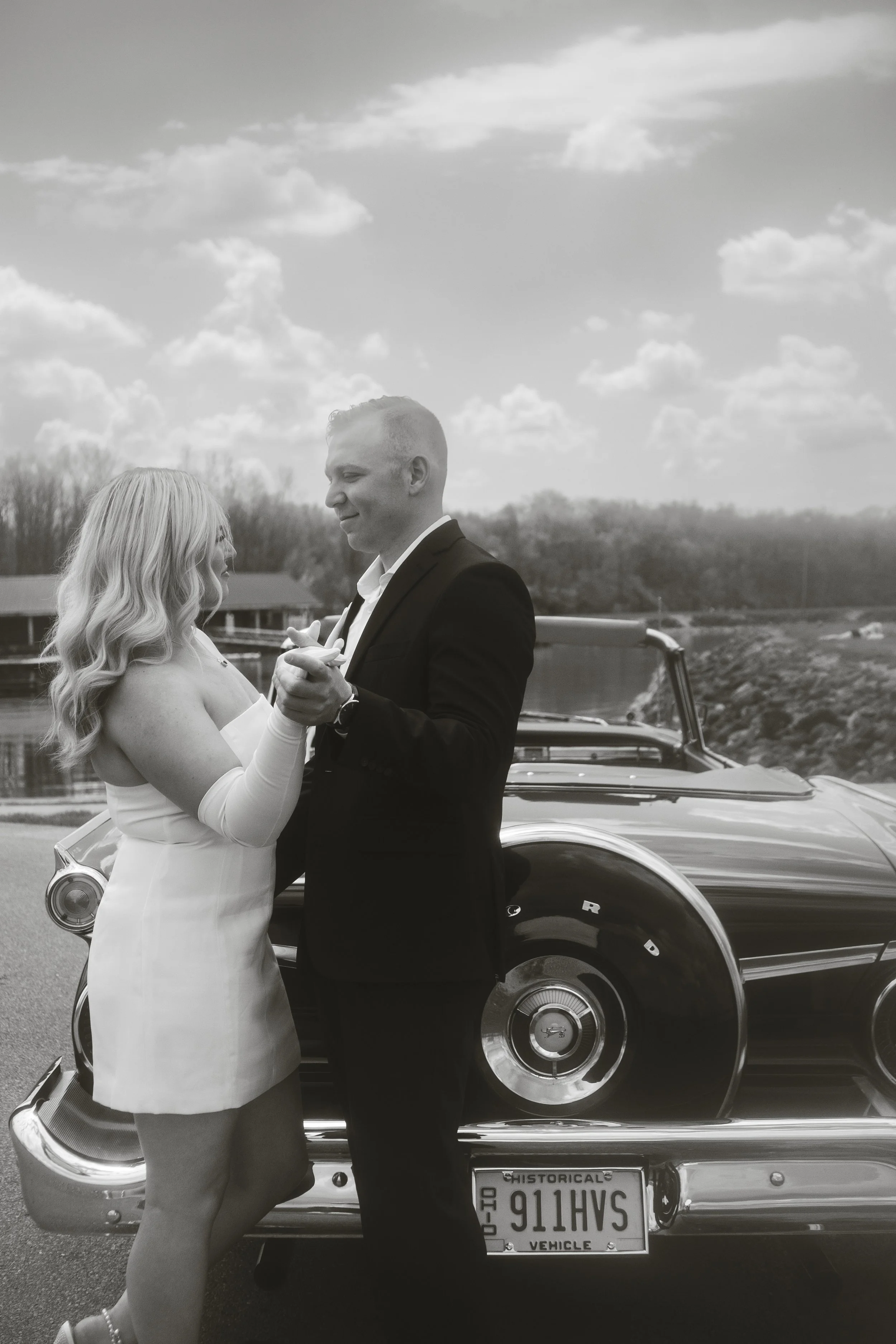 A black and white photo of a man and woman dancing in front of a vintage car by a body of water on a cloudy day.