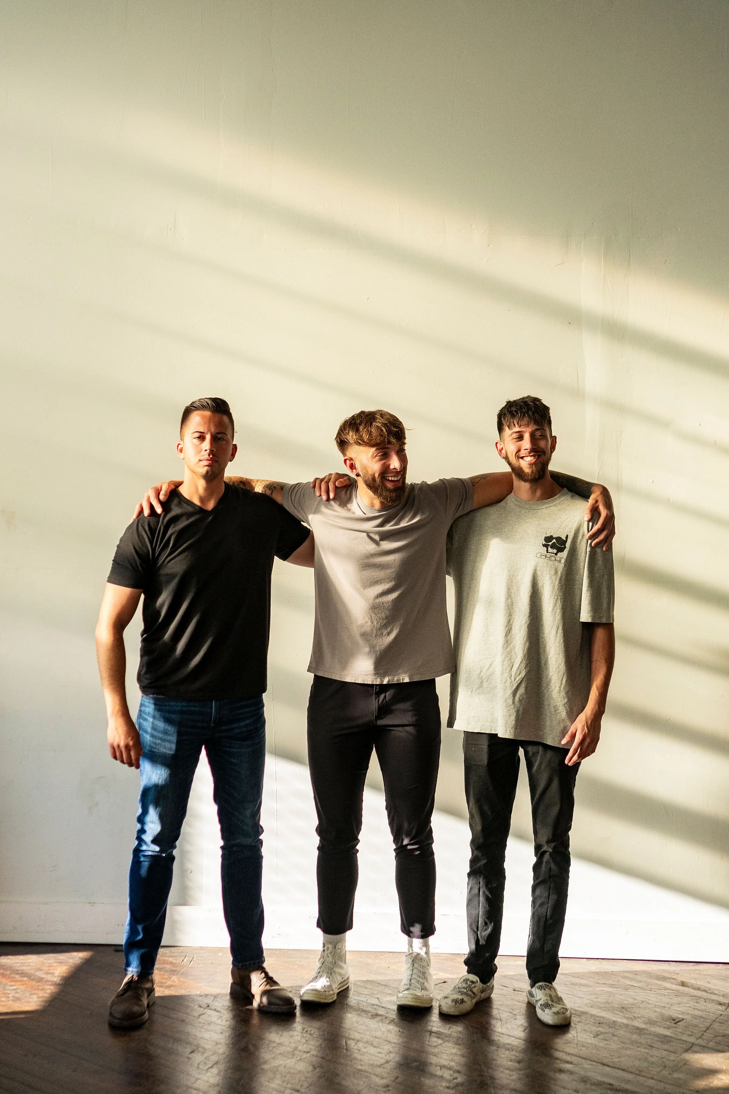 Three young men standing together, smiling, with their arms around each other in front of a plain wall with sunlight and shadows.