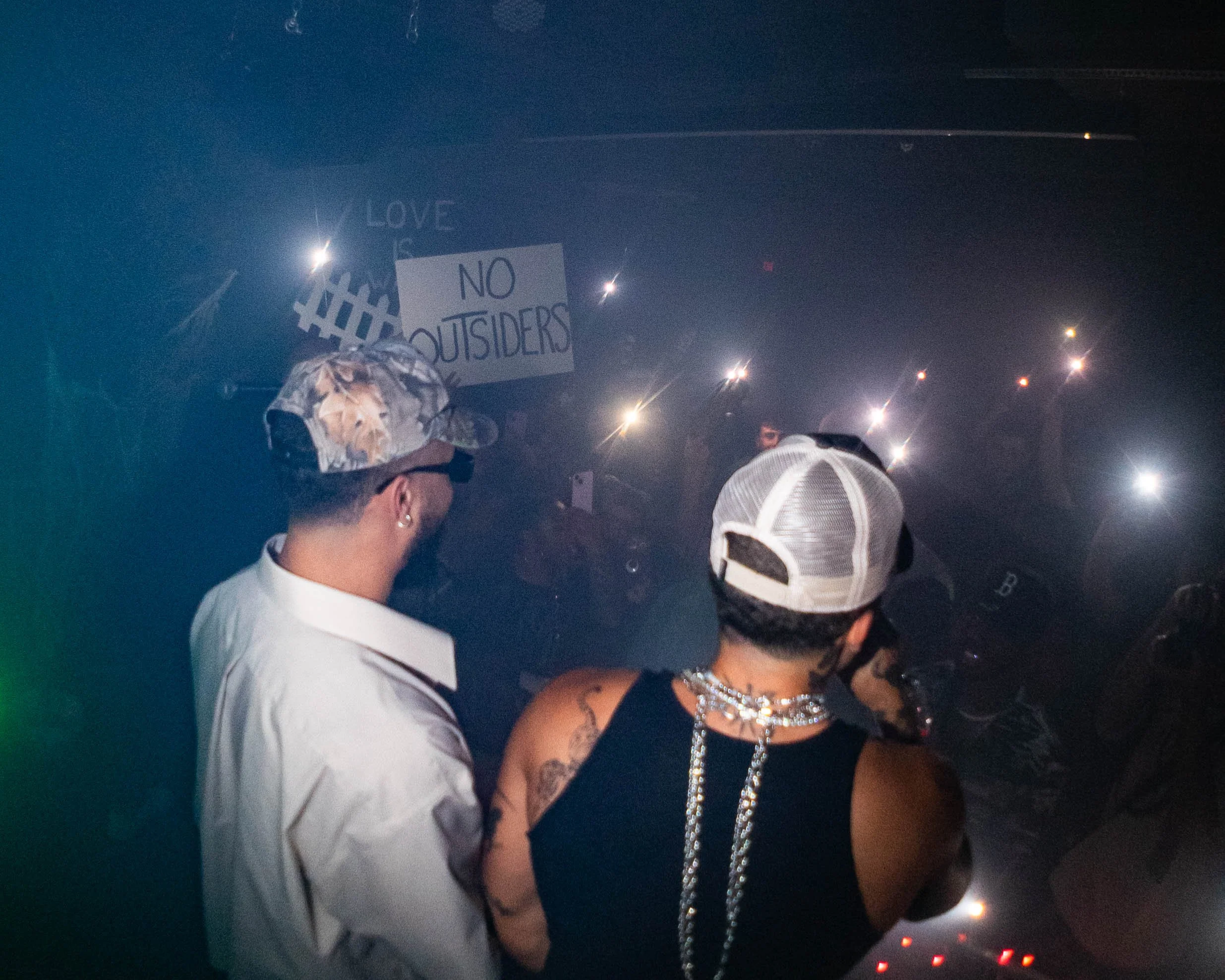 Two people with tattoos and wearing hats at a dark, smoky party with flashing lights. A sign in the background reads 'NO OUTSIDERS' and 'LOVE'.