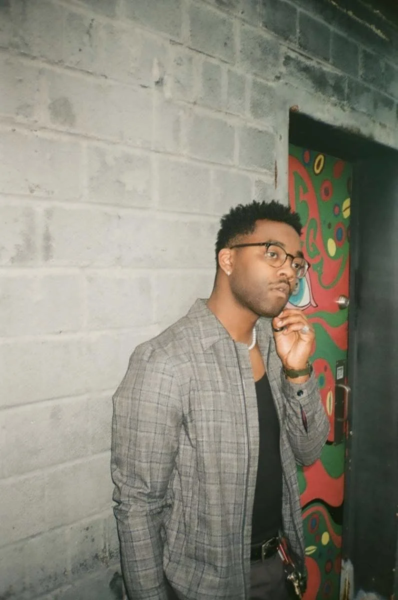 A young man with glasses, wearing a gray plaid blazer and black shirt, standing next to a colorful door with abstract designs, posing indoors against a concrete wall.
