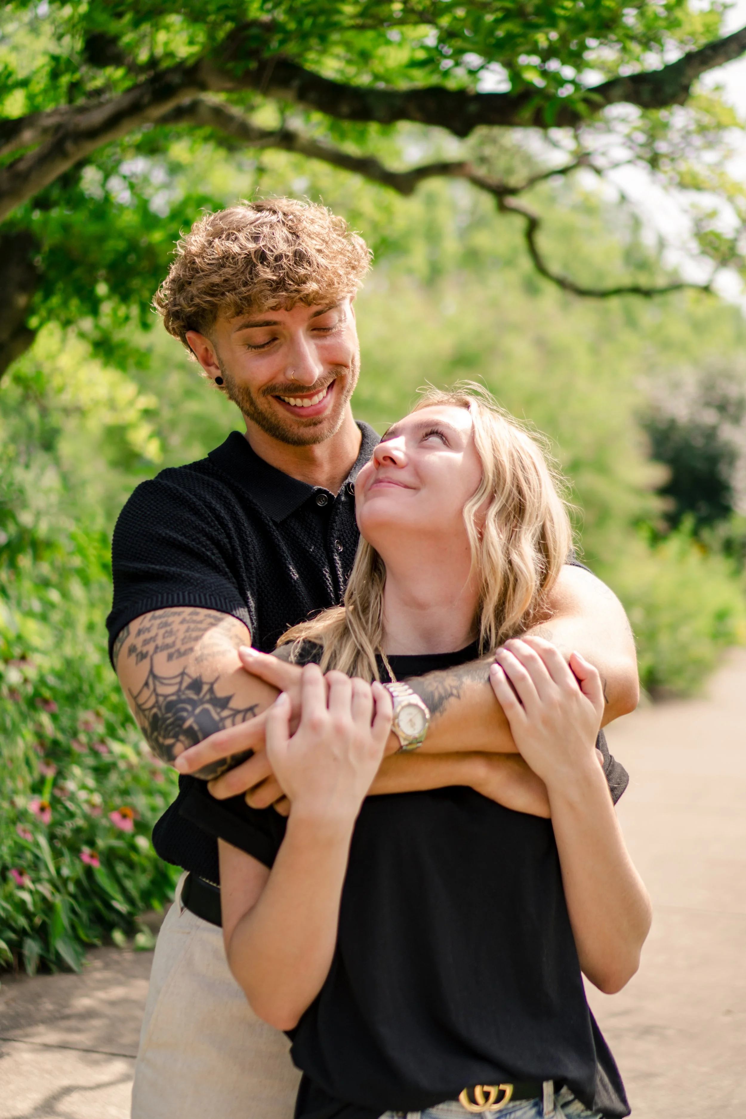 A young man with tattoos and a young woman sharing a hug outdoors on a sunny day, with green trees and flowers in the background.