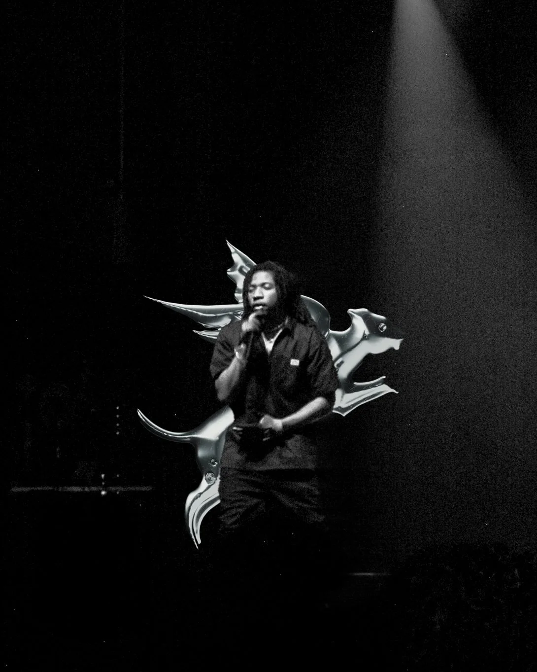 A performer with dreadlocks singing into a microphone against a dark background with abstract metallic shapes.