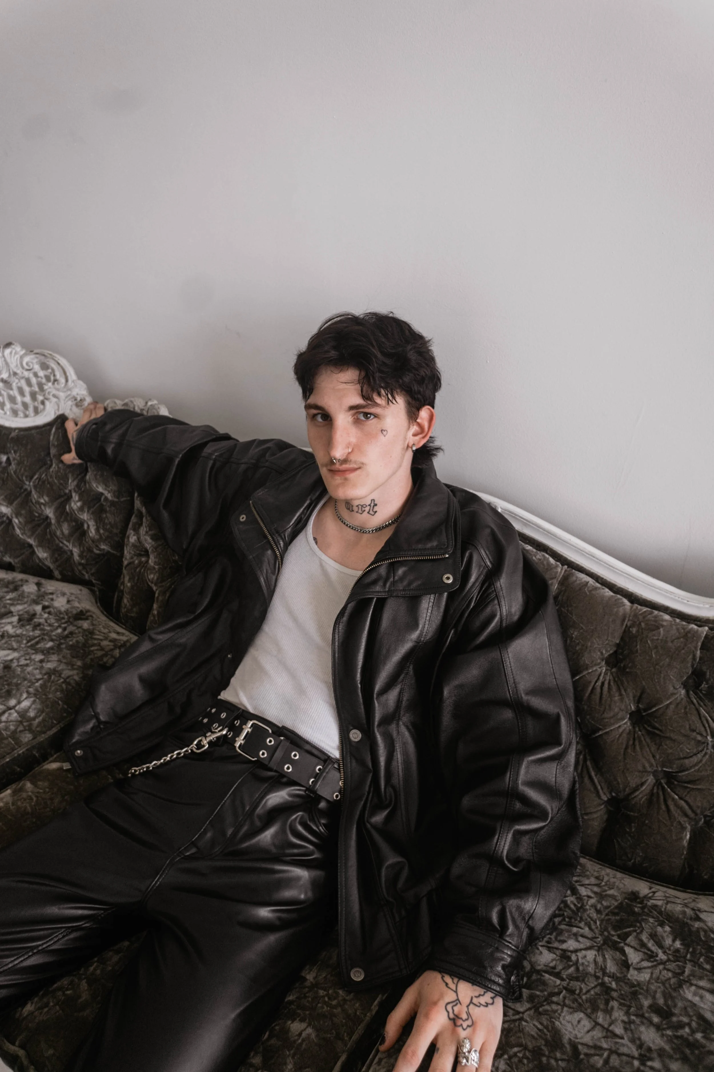 A young man with dark hair sitting on a vintage, ornate, dark velvet sofa, wearing black leather pants, a black leather jacket, and a white tank top, with tattoos, jewelry, and makeup.