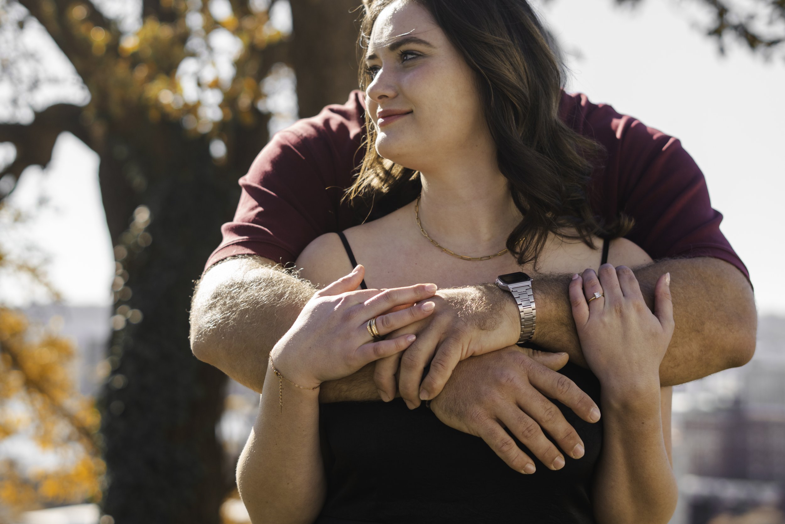 A woman with brown hair and a black top is outdoors, while a man with a gold watch and a red shirt embraces her from behind, wrapping their arms around her shoulders and waist. The woman looks to her right with a slight smile.