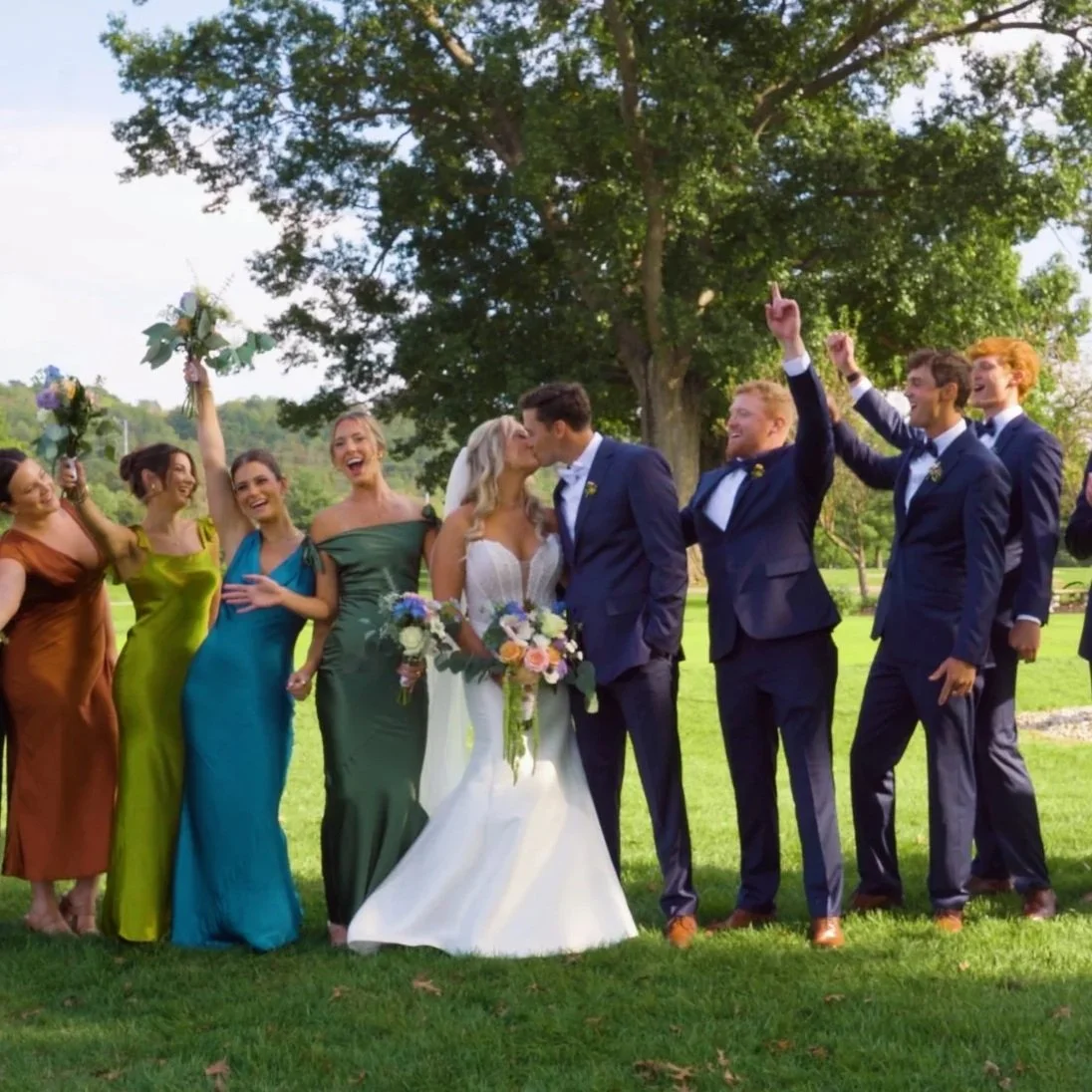 A wedding celebration in a park with the bride and groom kissing, surrounded by friends in colorful dresses and suits, some celebrating with raised fists and smiles, under a large tree.