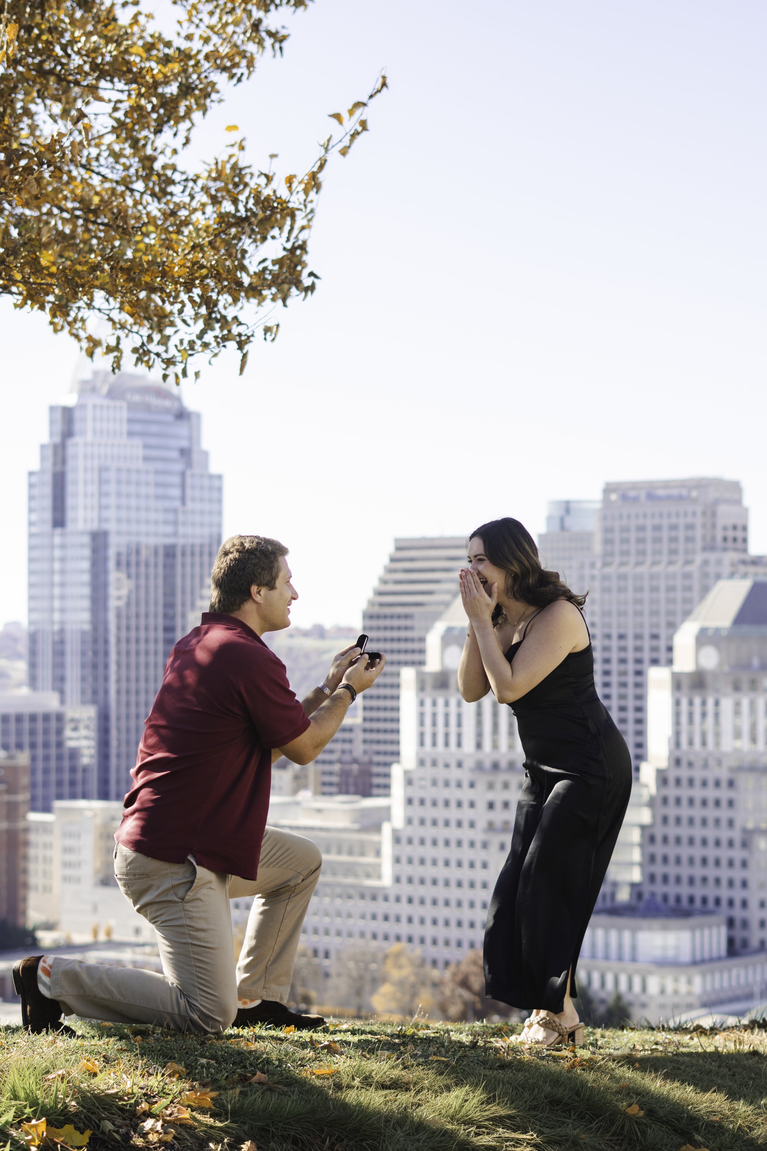 A man on one knee proposing to a woman in an outdoor park with city buildings in the background.