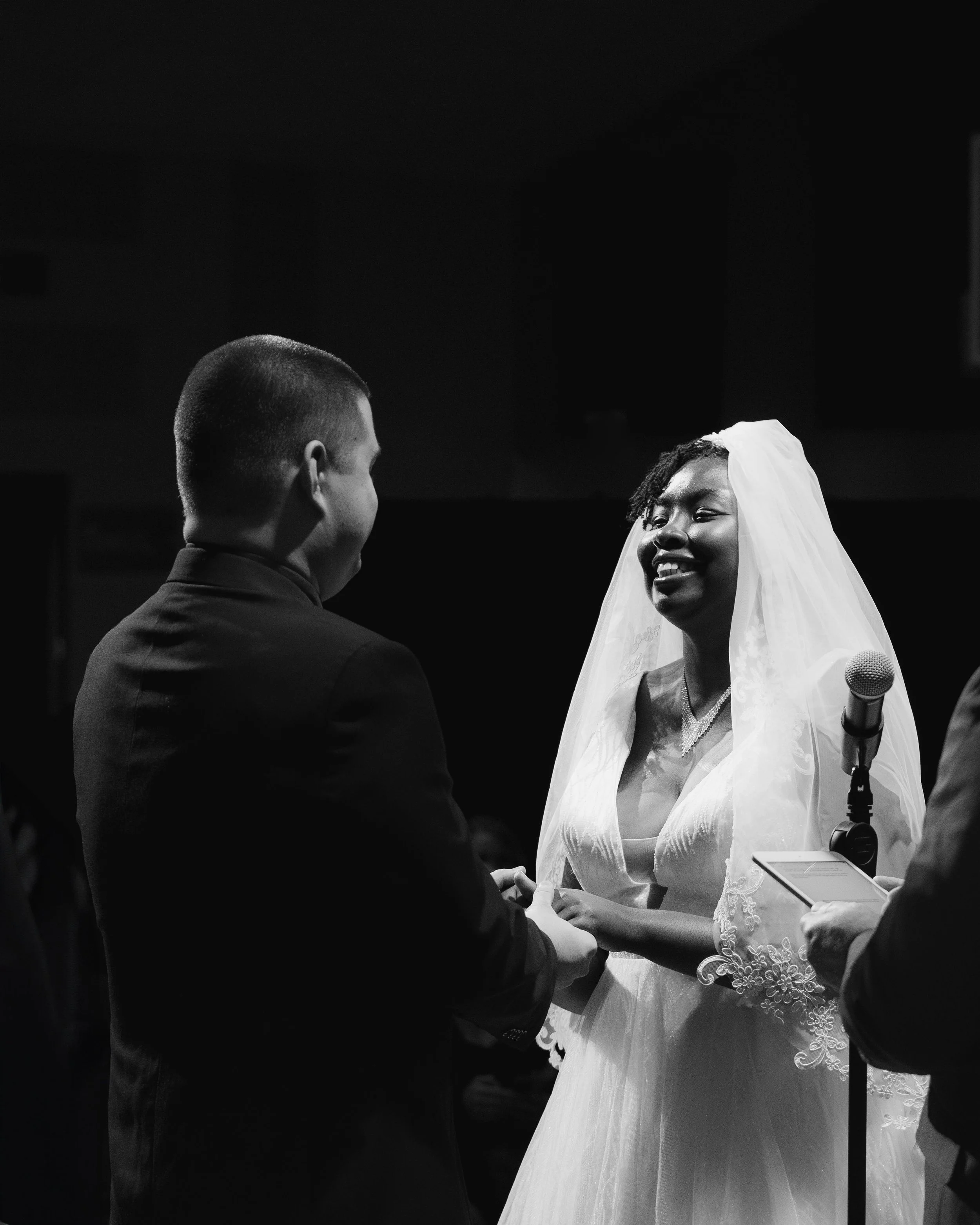 Black and white photo of a bride and groom holding hands during a wedding ceremony, with the officiant holding a tablet nearby.