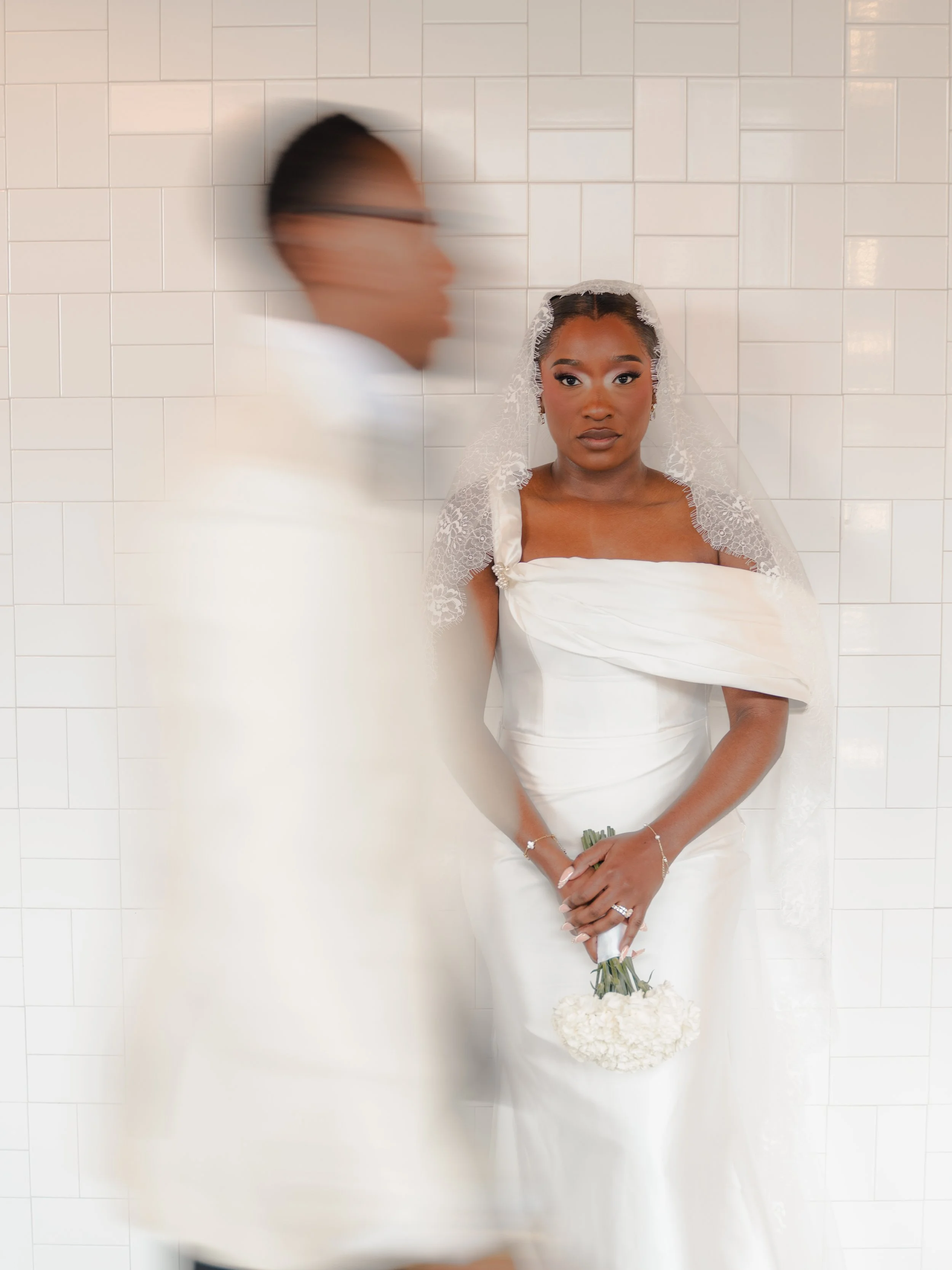 A bride in a white wedding dress and lace veil holding a bouquet of white flowers, standing against a tiled white wall. A man in a white suit, slightly blurred, stands next to her.