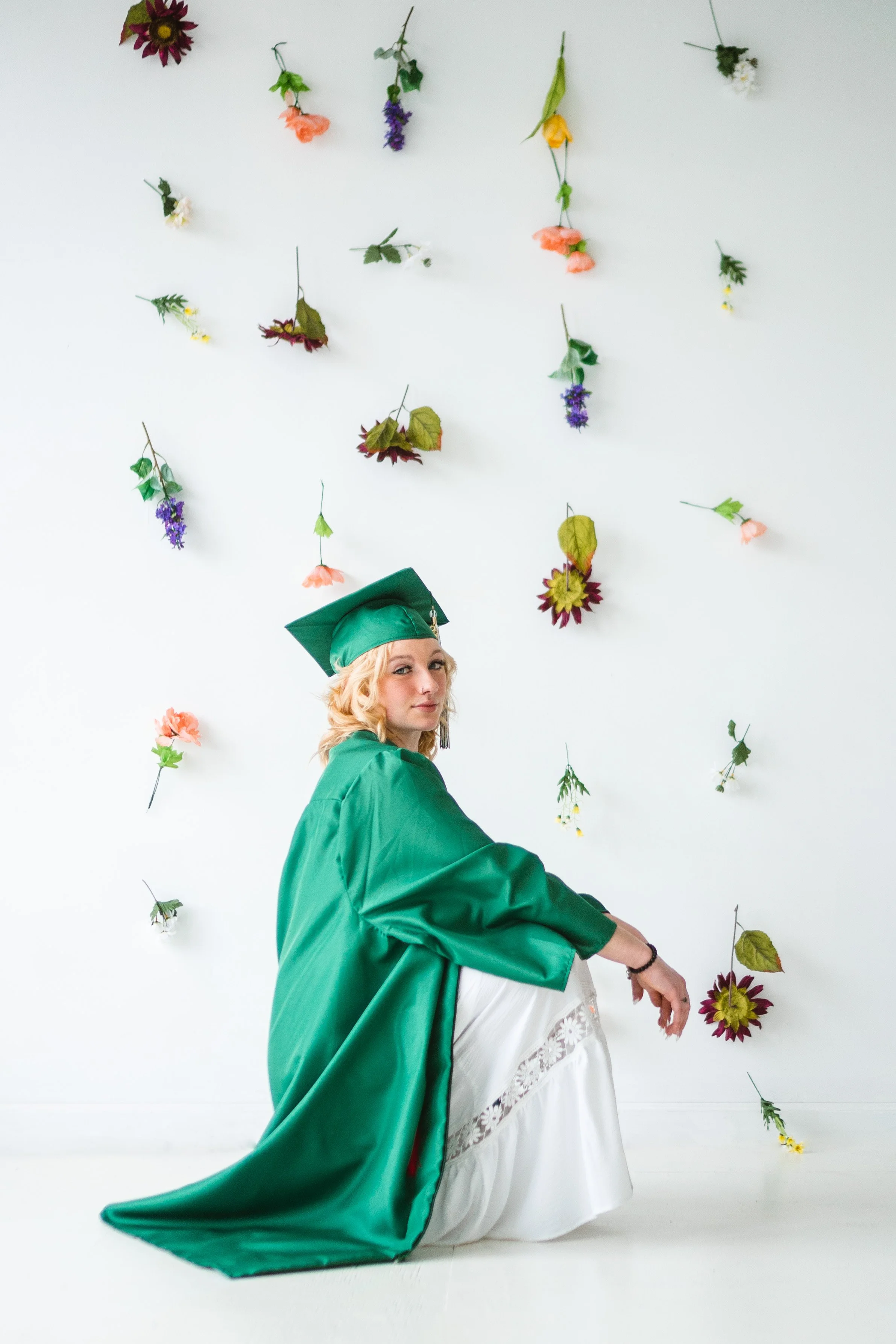 A young woman in a green graduation gown and cap sitting on the floor in front of a white wall decorated with hanging colorful flowers.