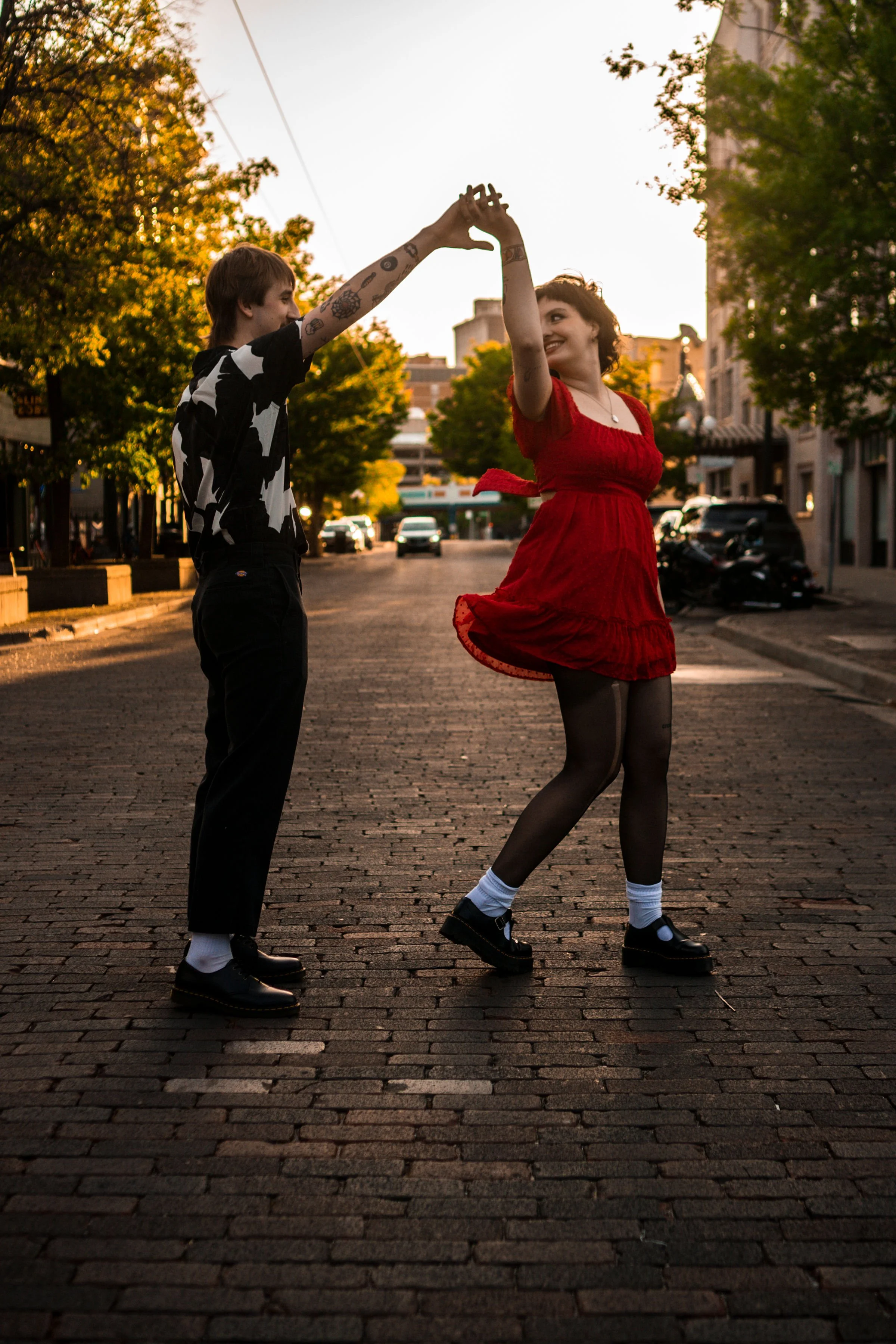 A young man and woman dancing on a cobblestone street at sunset, holding hands and smiling, with trees and buildings in the background.
