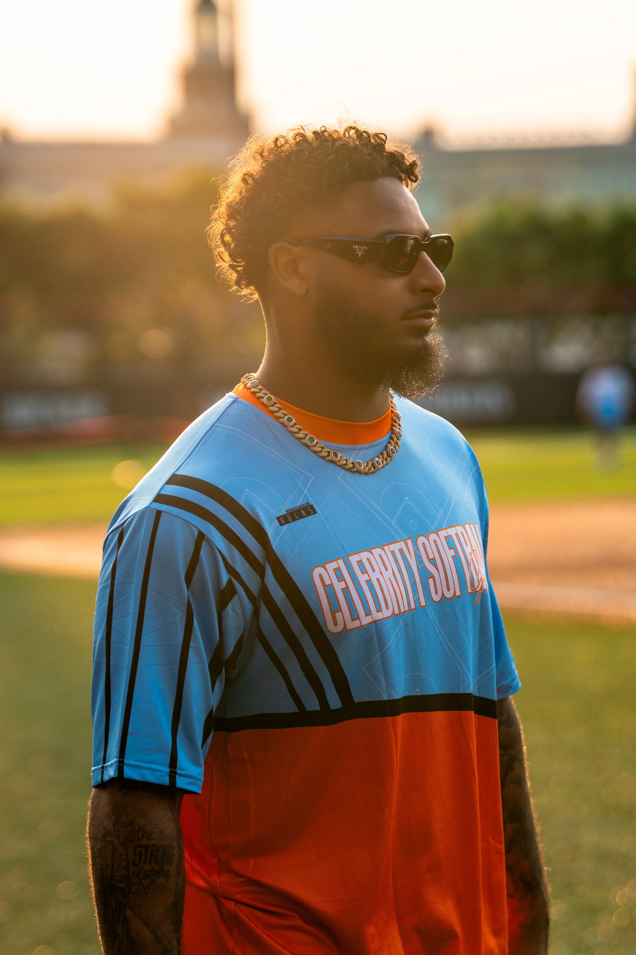 A young man with curly hair wearing sunglasses, a colorful graphic T-shirt, and a shell necklace, standing outdoors in the late afternoon sunlight.