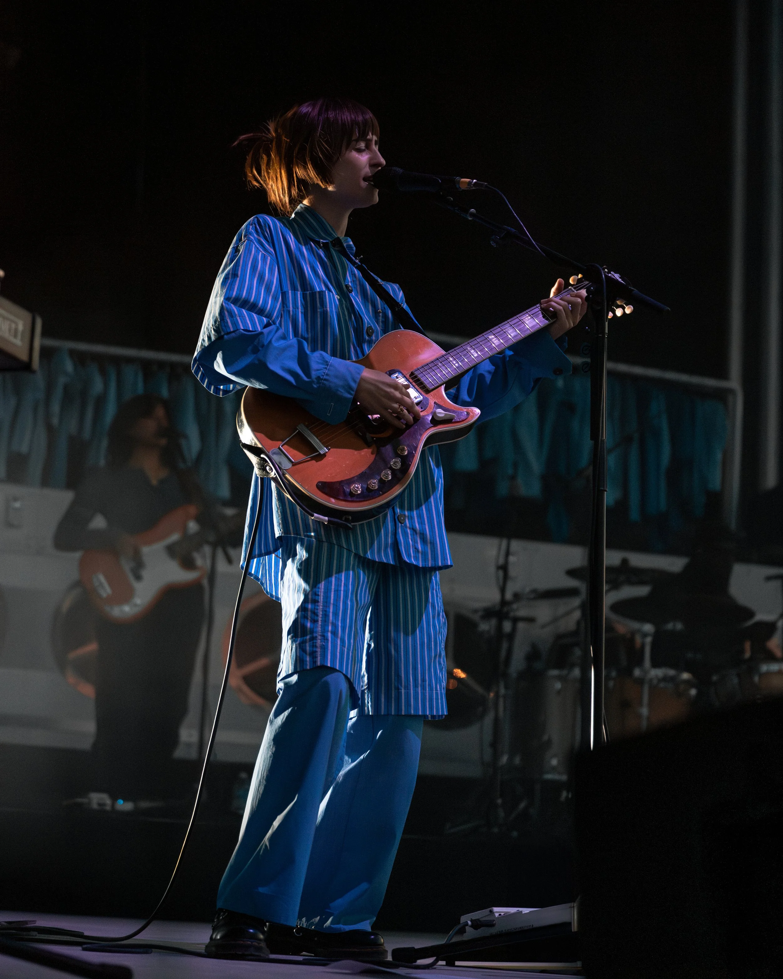 A woman with short brown hair in striped blue pajamas playing an electric guitar and singing on stage, with a woman in the background also playing guitar.