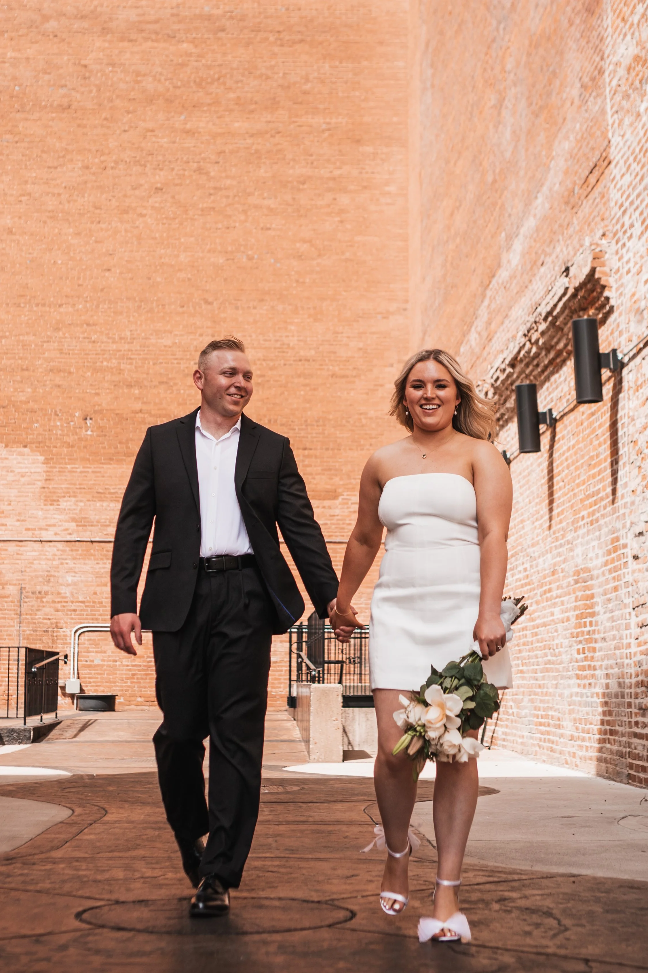 A smiling couple in wedding attire holding hands while walking outdoors against a brick wall background. The woman is wearing a white strapless dress and heels, carrying a bouquet of flowers. The man is dressed in a black suit and white shirt.