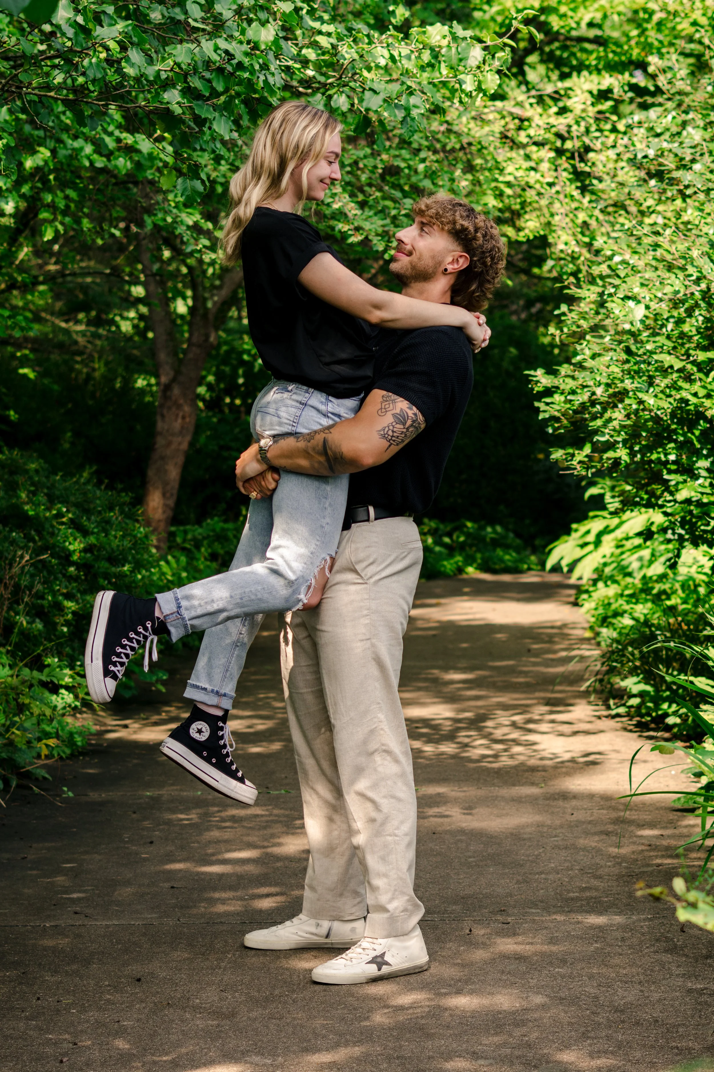 A man lifting a woman in his arms on a wooded path, both smiling and looking at each other amidst greenery.