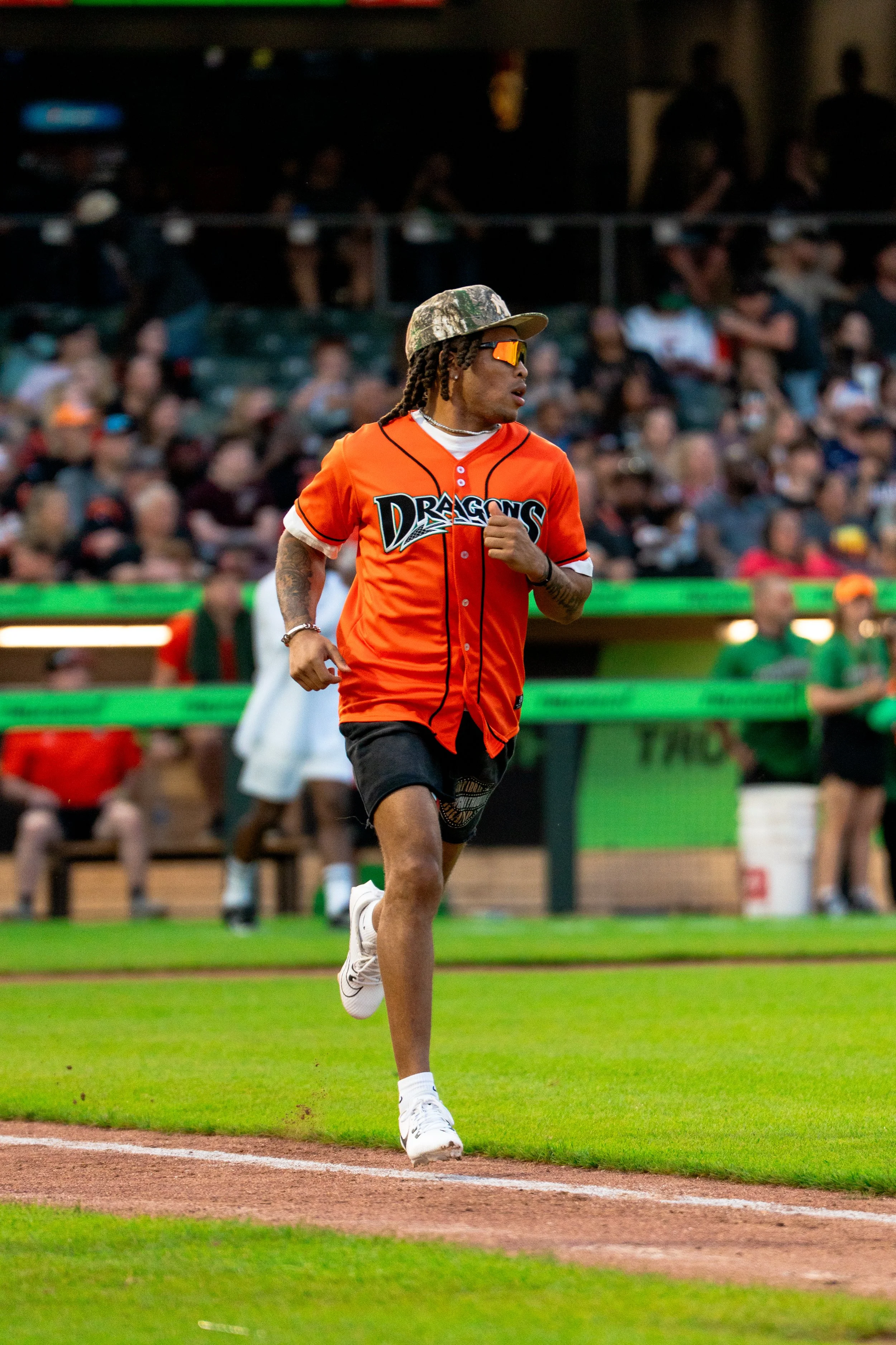 A man running on a baseball field, wearing an orange jersey, black shorts, a camouflage cap, sunglasses, and jewelry, with people in the background.