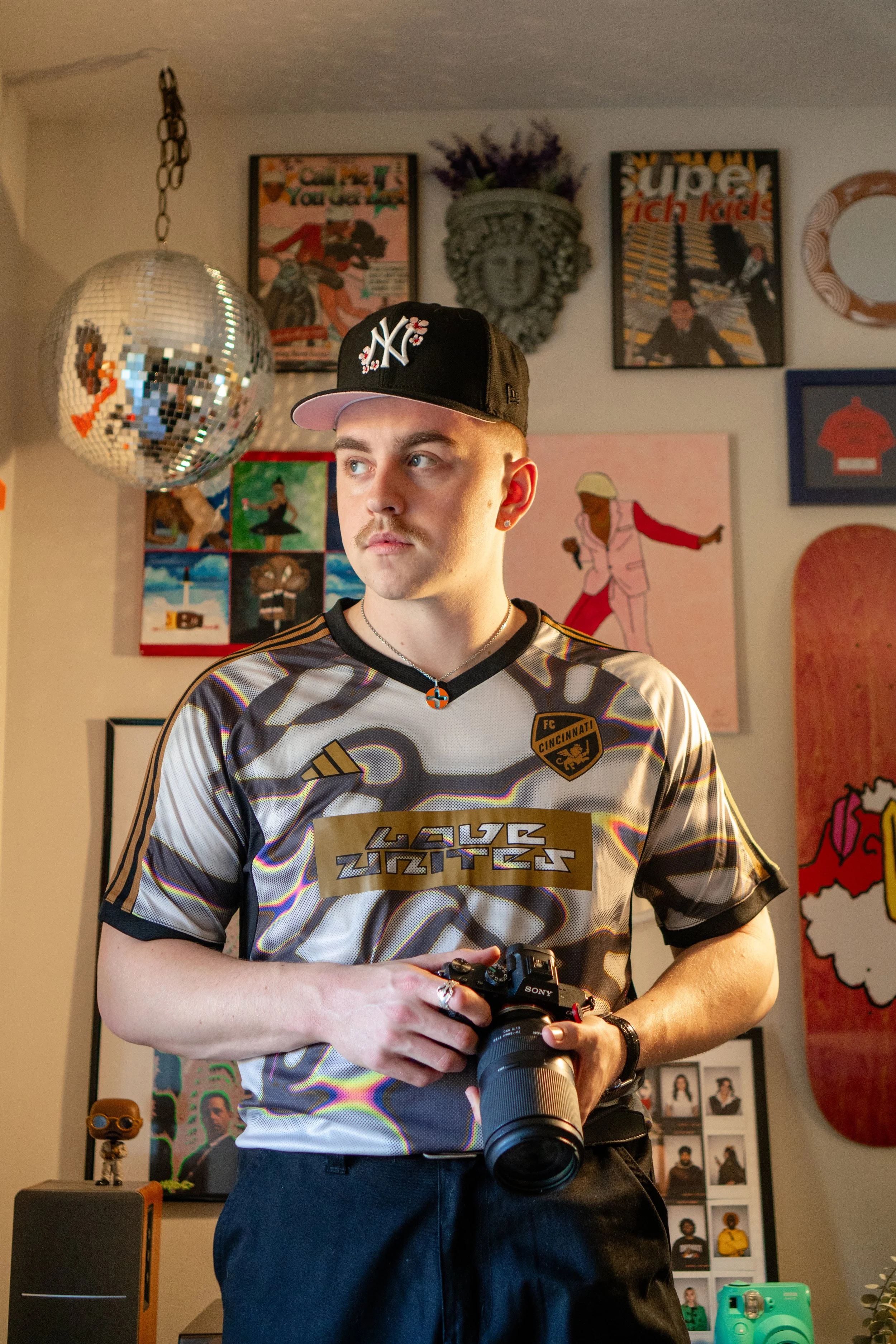 Young man wearing a New York Yankees cap and a FC Cincinnati soccer jersey, holding a Sony camera, standing in a art-filled room with posters, paintings, and decor on the walls.