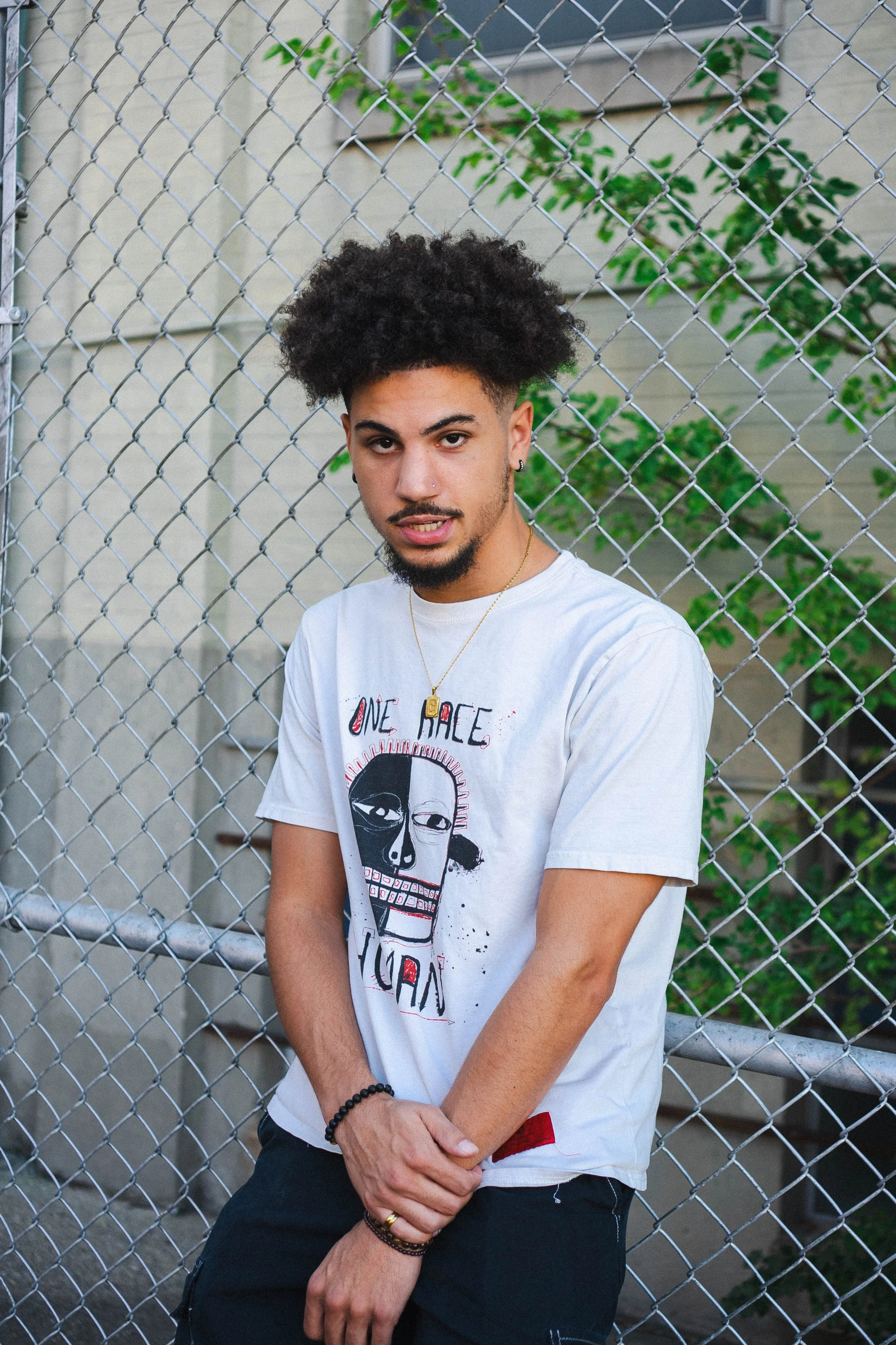 A young man with curly hair and a beard stands in front of a chain-link fence with green foliage behind him. He wears a white graphic t-shirt and jewelry.