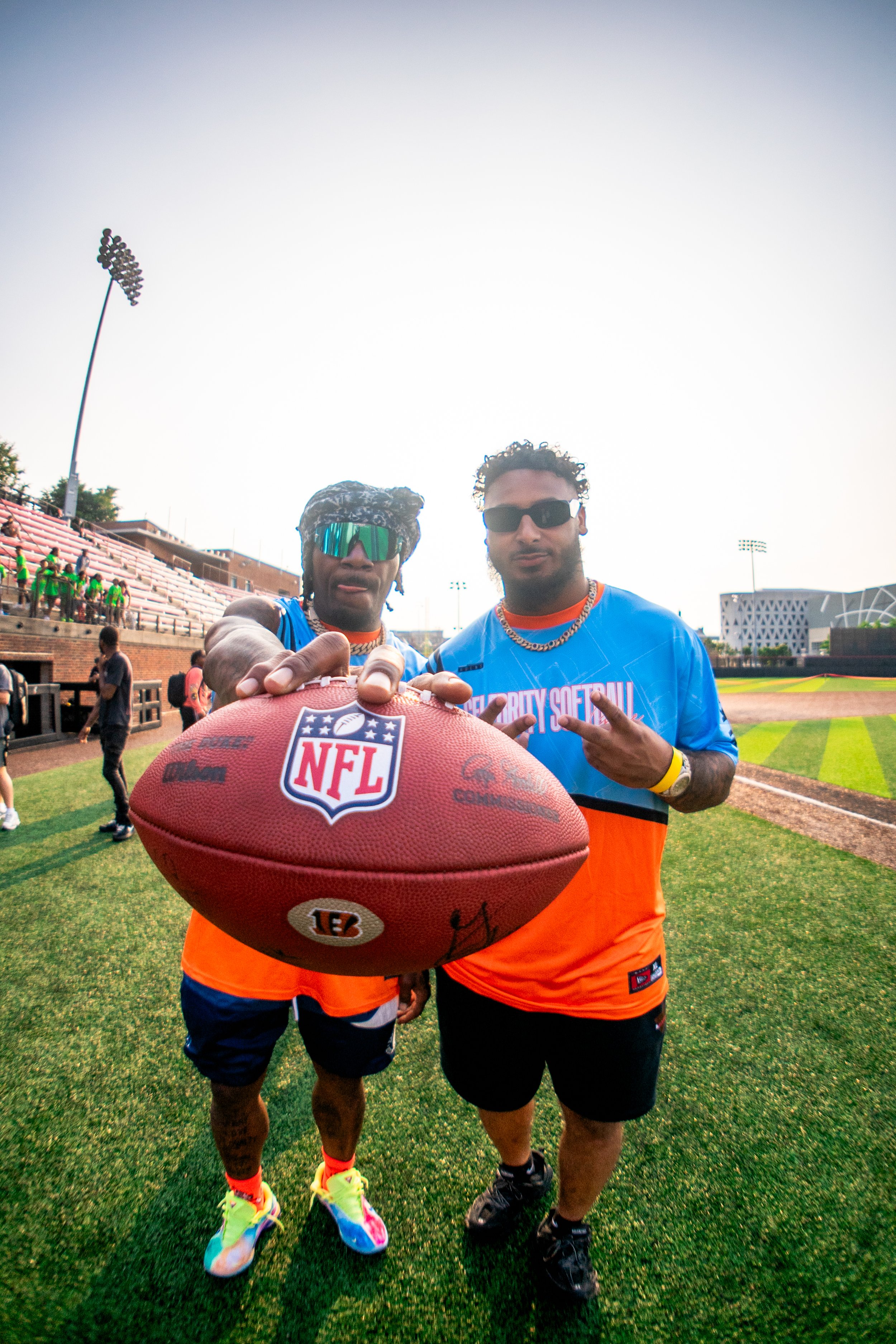 Two men standing on a football field, holding an NFL football, with a stadium and people in the background. Both are wearing sunglasses and sportswear.