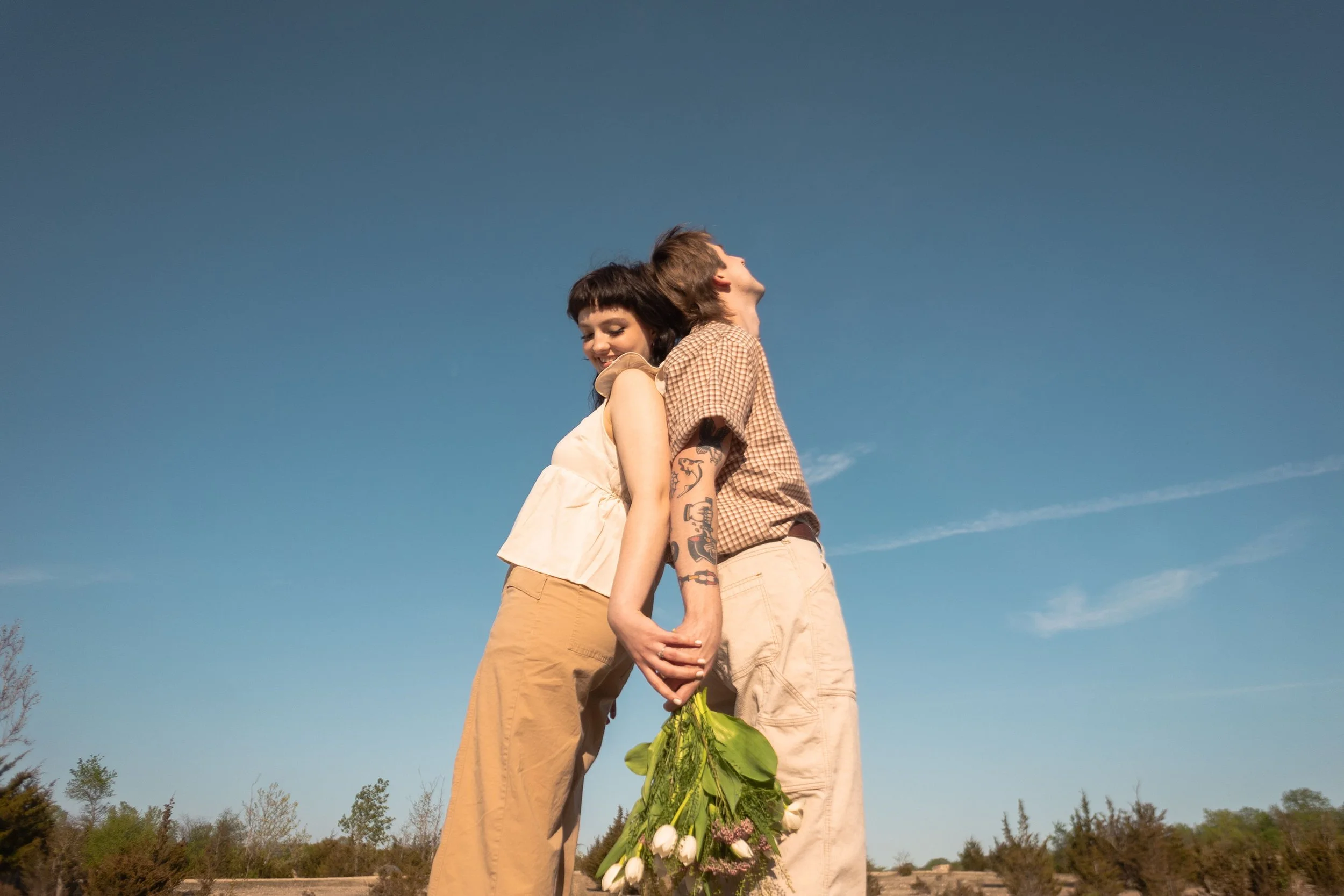 A young man and woman holding hands, standing back to back outside under a clear blue sky, with the woman holding a bouquet of white tulips and purple flowers.