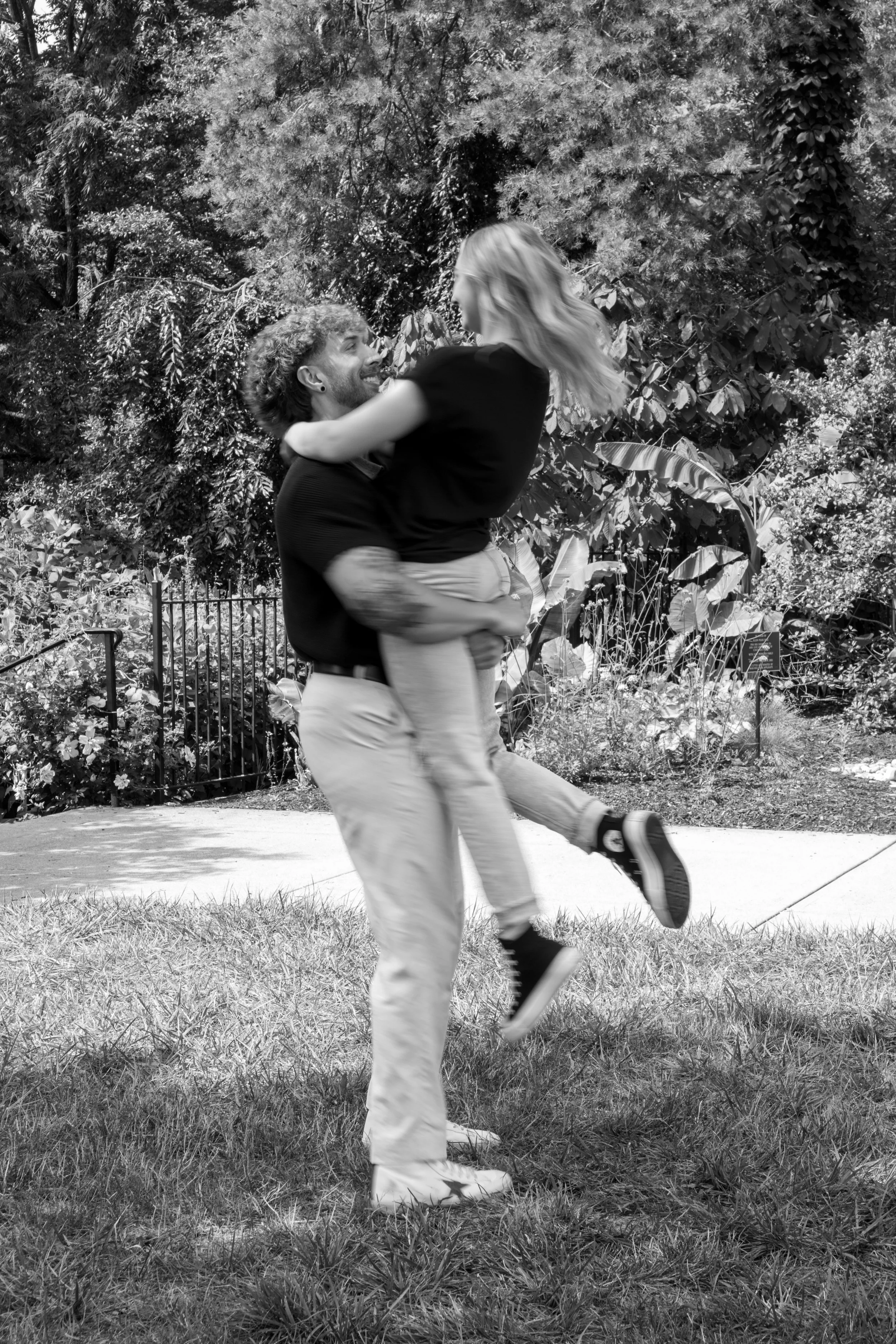 A happy man lifting and holding a woman in an outdoor park or garden setting, with trees and bushes in the background.