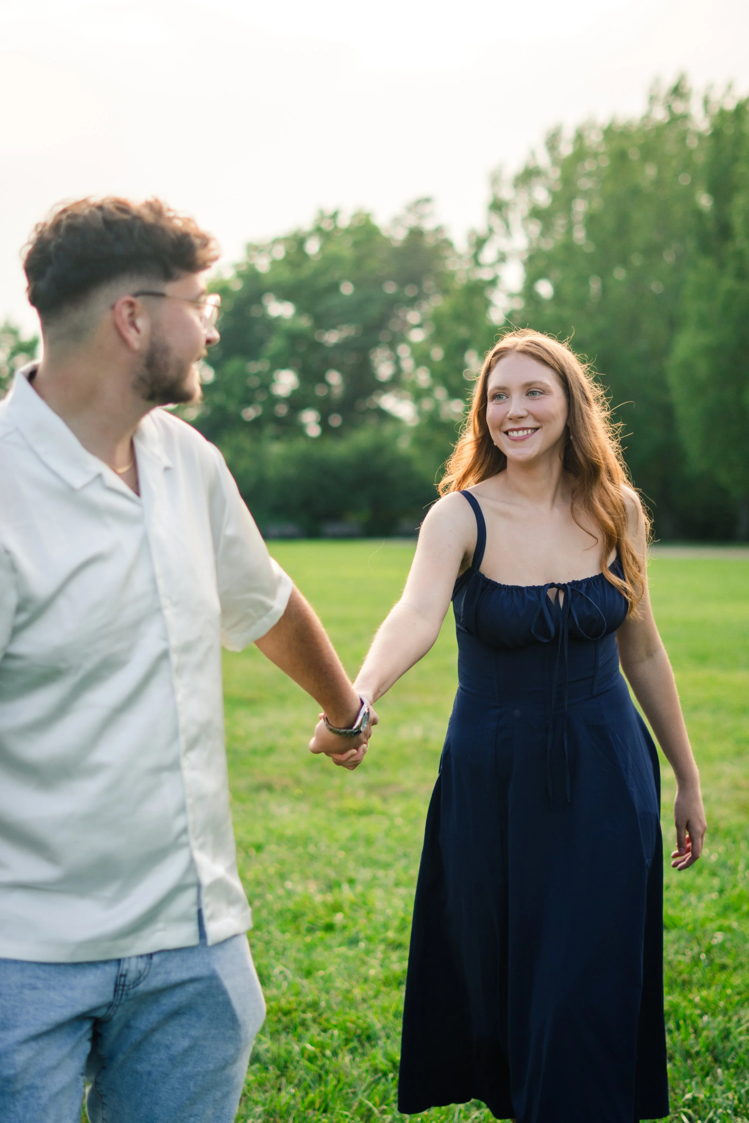 A young man and woman holding hands in a grassy park, with trees in the background, enjoying a sunny day.