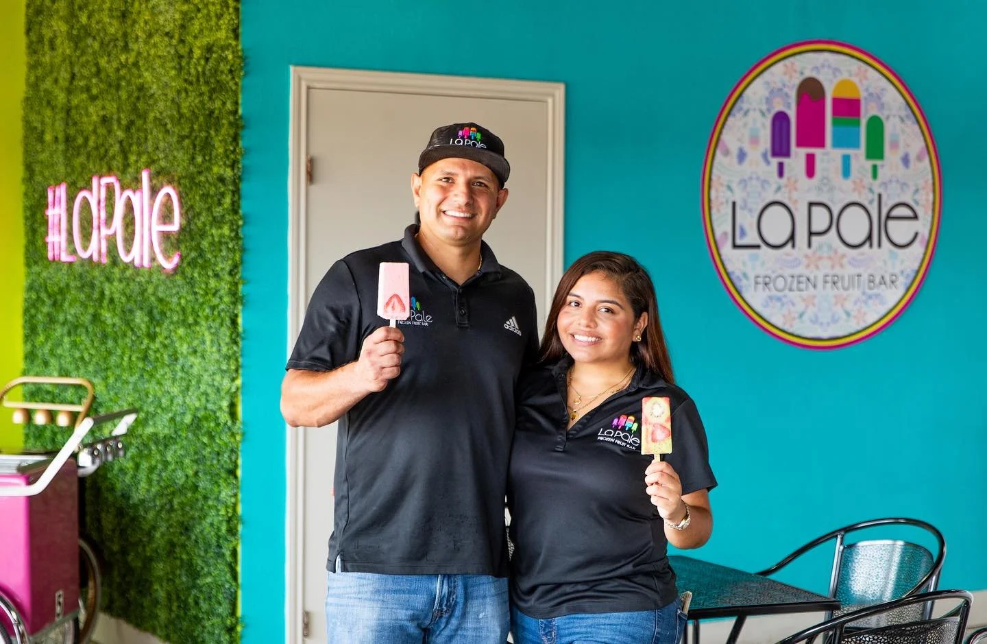 Two smiling employees in black polo shirts with 'La Pale' logo, holding popsicles inside a colorful frozen fruit bar shop.