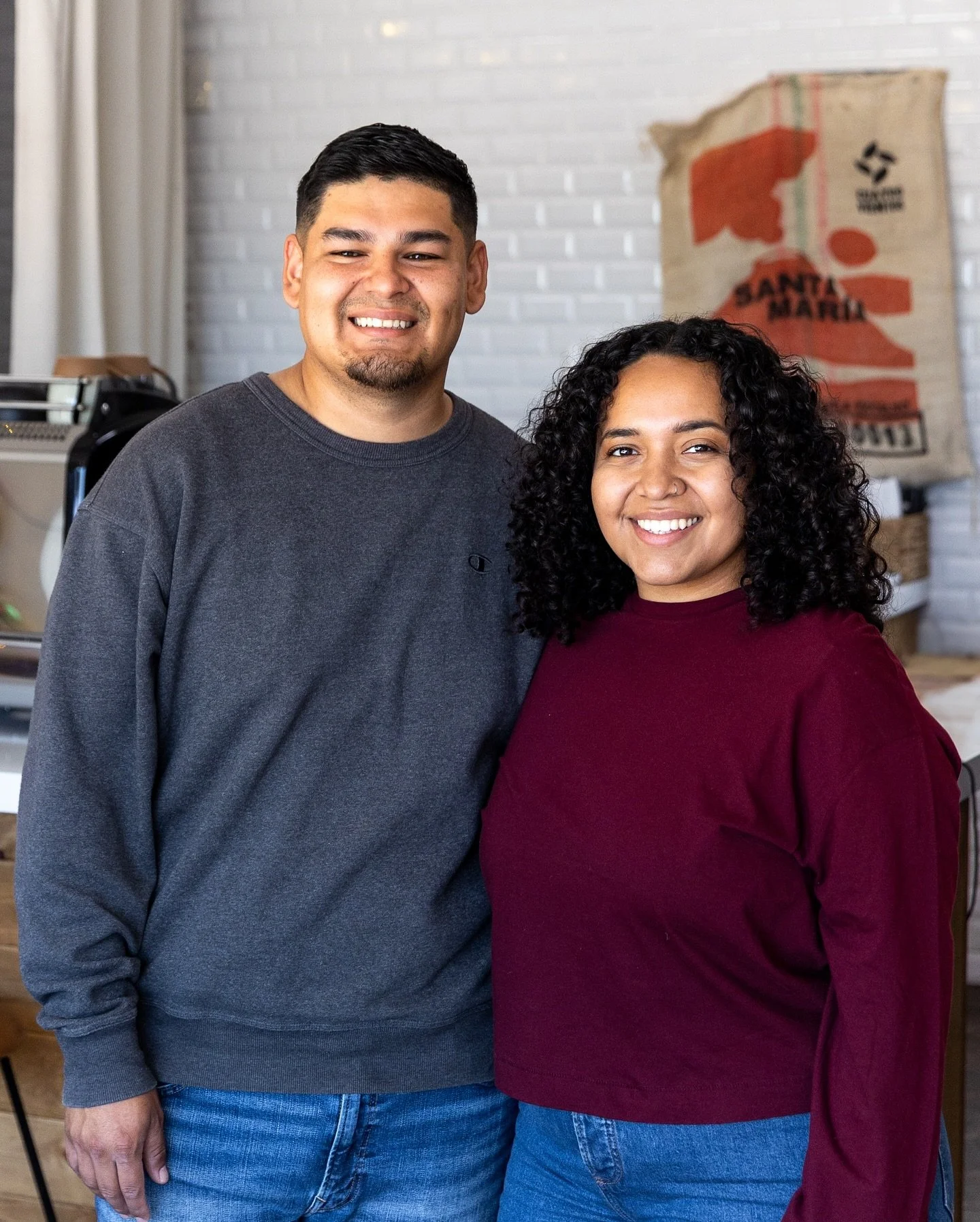 A man and a woman smiling inside a room with white brick wall background. The man is wearing a dark gray long-sleeve shirt and jeans, and the woman is wearing a maroon long-sleeve shirt and jeans. 