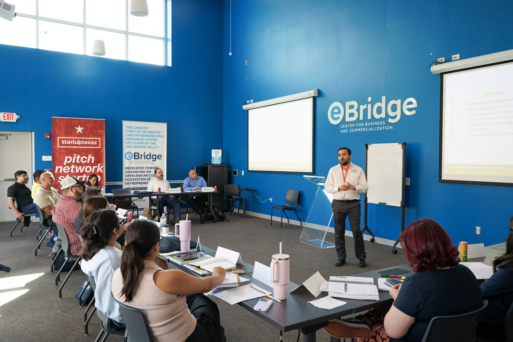 A man standing in front of a blue wall with the eBridge logo giving a presentation to seated attendees in a conference room. The room has large windows, two projection screens, and banners related to startups and entrepreneurship.