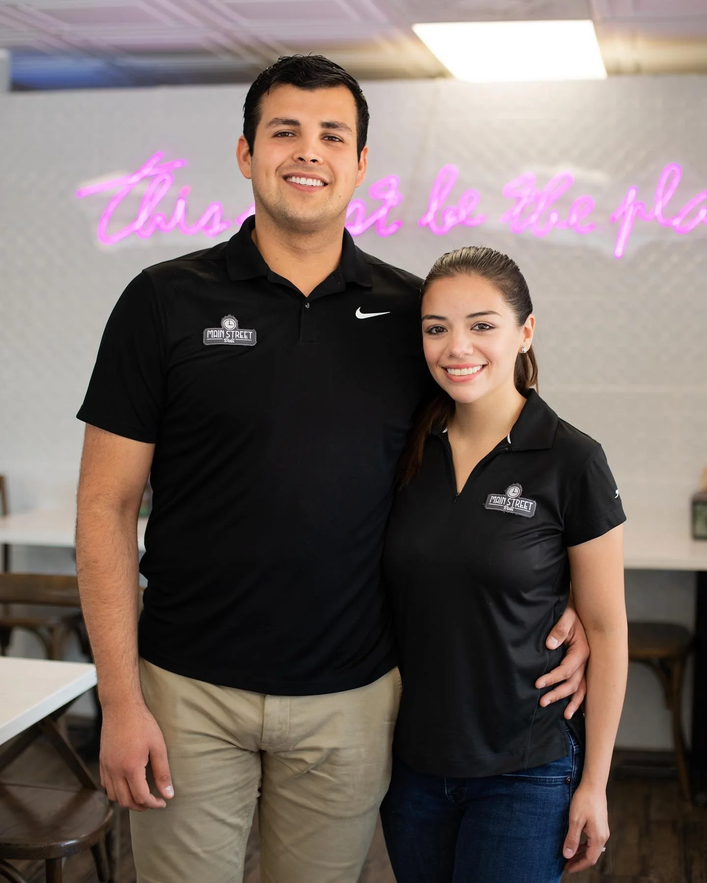 A man and a woman smiling and standing together inside a restaurant or cafe, both wearing black shirts with a 'Main Street' logo. The background features a wall with a purple neon sign that reads 'this or be the p...'.