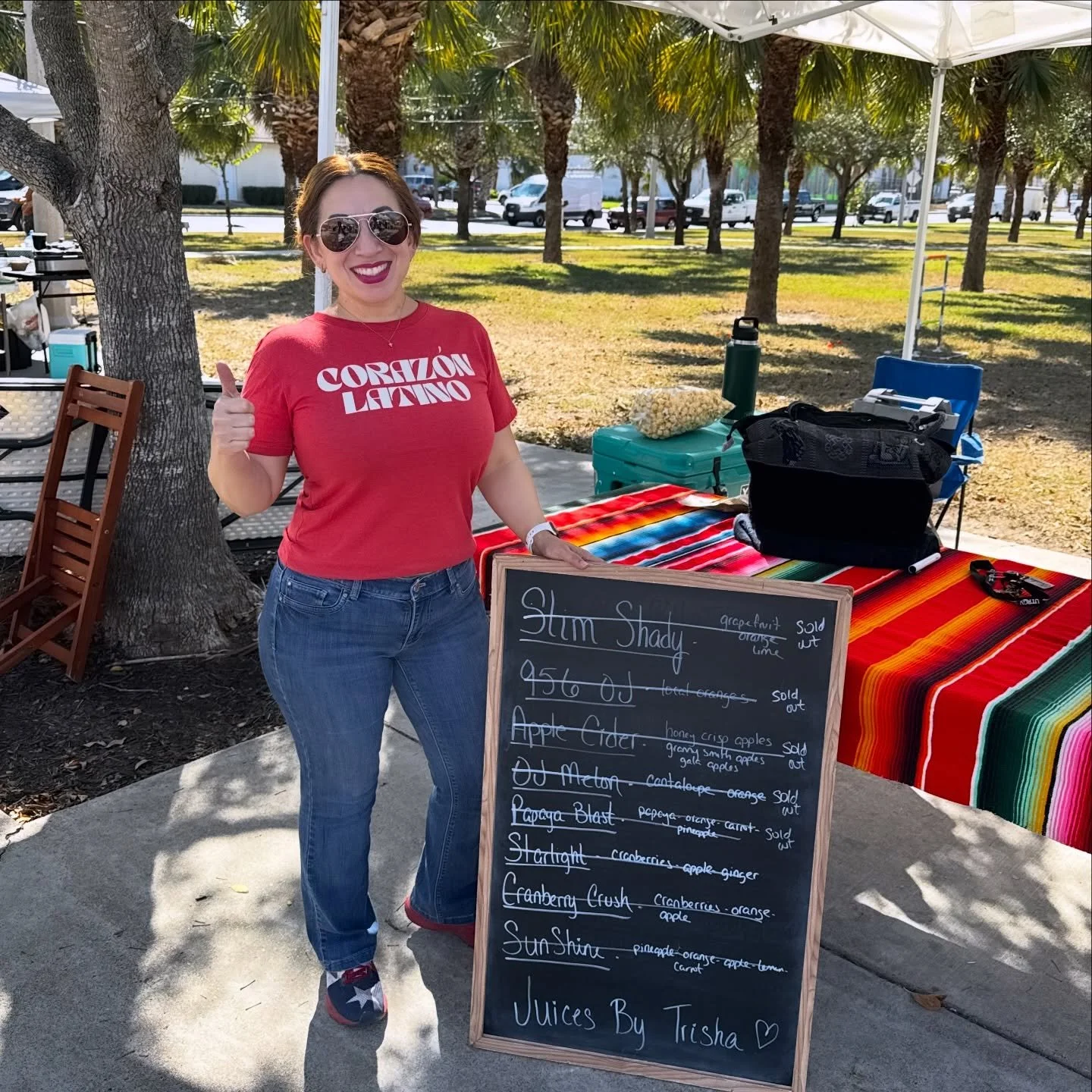 Woman standing outdoors next to a blackboard sign with juice menu, giving a thumbs up, wearing sunglasses and a red t-shirt, underneath a white canopy with a striped tablecloth behind her.
