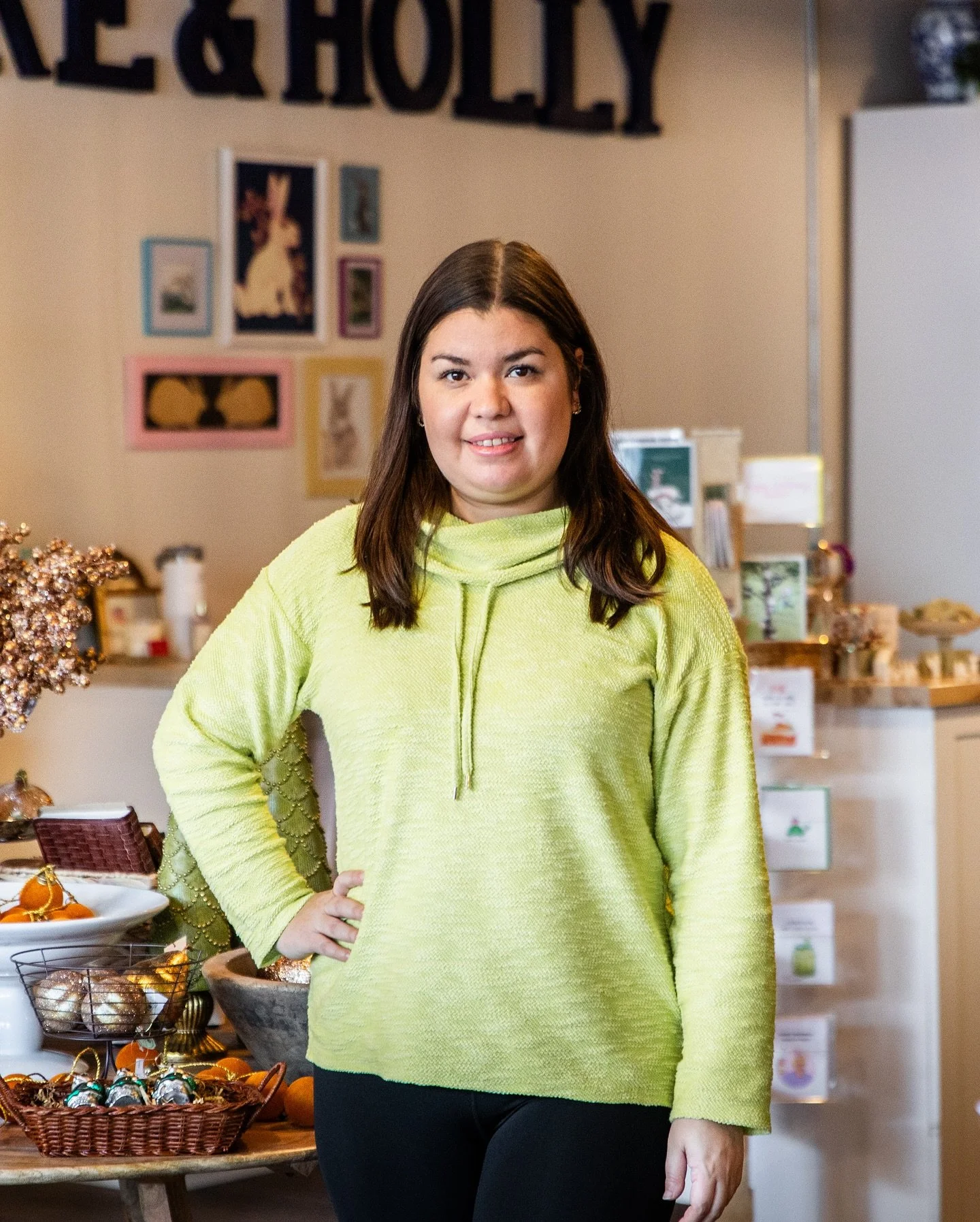 A young woman with shoulder-length dark hair, wearing a lime green sweater, standing indoors in front of a decorated wall with framed pictures and a sign that says 'PEACE & HOLY.' She is smiling and has one hand on her hip. There are decorative items and a basket of ornaments on a table to her left.