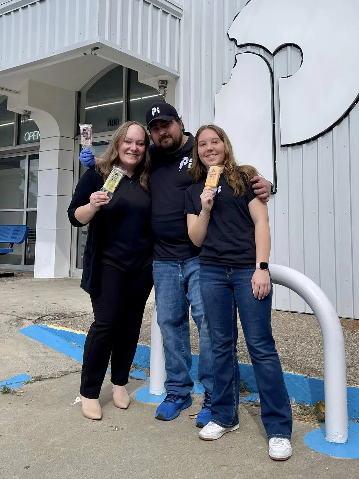 Three people standing outside a building, holding ice cream bars, smiling, and posing for a photo.