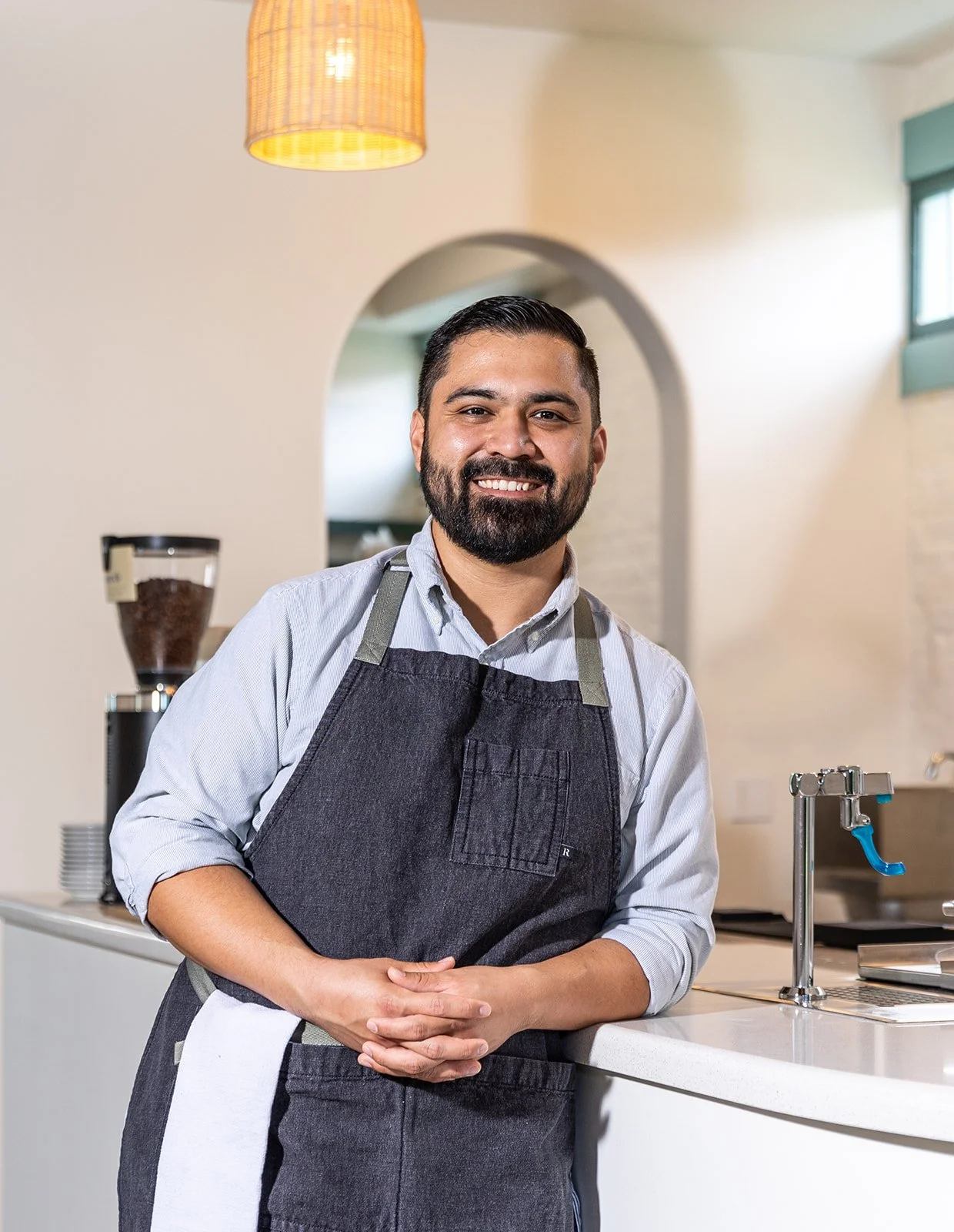 Smiling man with a beard wearing a gray apron standing behind a kitchen counter in a cafe or restaurant.