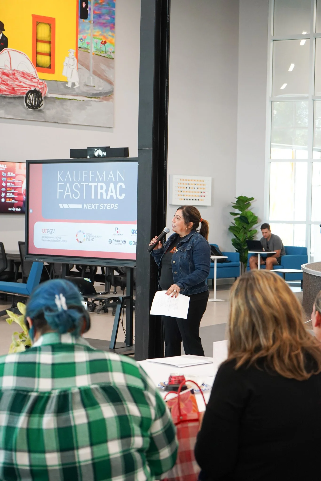 A woman is speaking into a microphone while holding papers during a presentation at a conference. A screen next to her displays the text 'KAUFFMAN FAST TRAC NEXT STEPS.' Several people are seated, facing the speaker, with some taking notes or listening attentively.