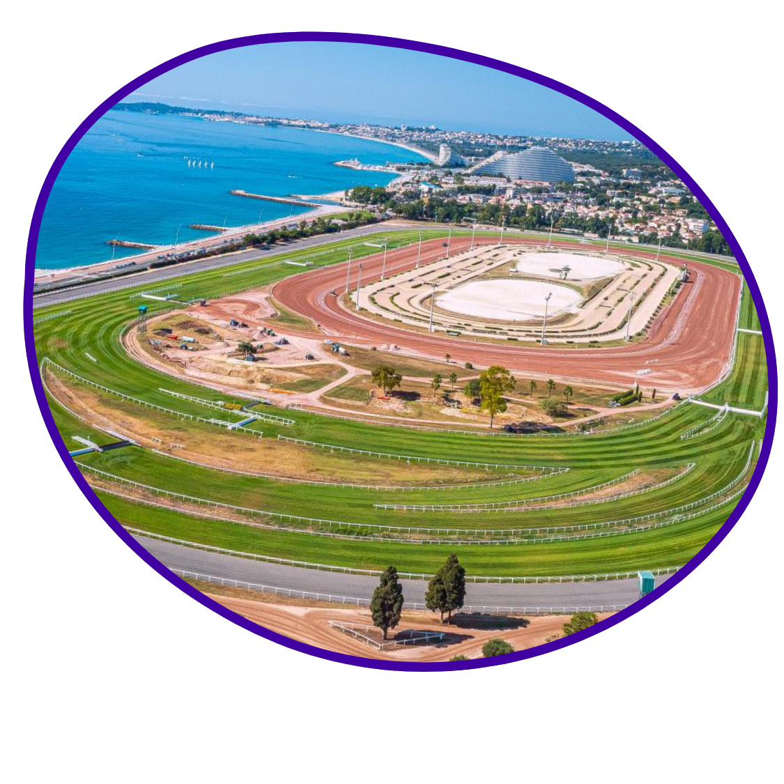 Une vue aérienne de l'hippodrome Côte d'Azur à Cagnes sur mer, avec la piste en terre rouge, les stands et le paysage urbain et maritime en arrière-plan. Une incroyable piste pour courir avec tes amis et les enfants.