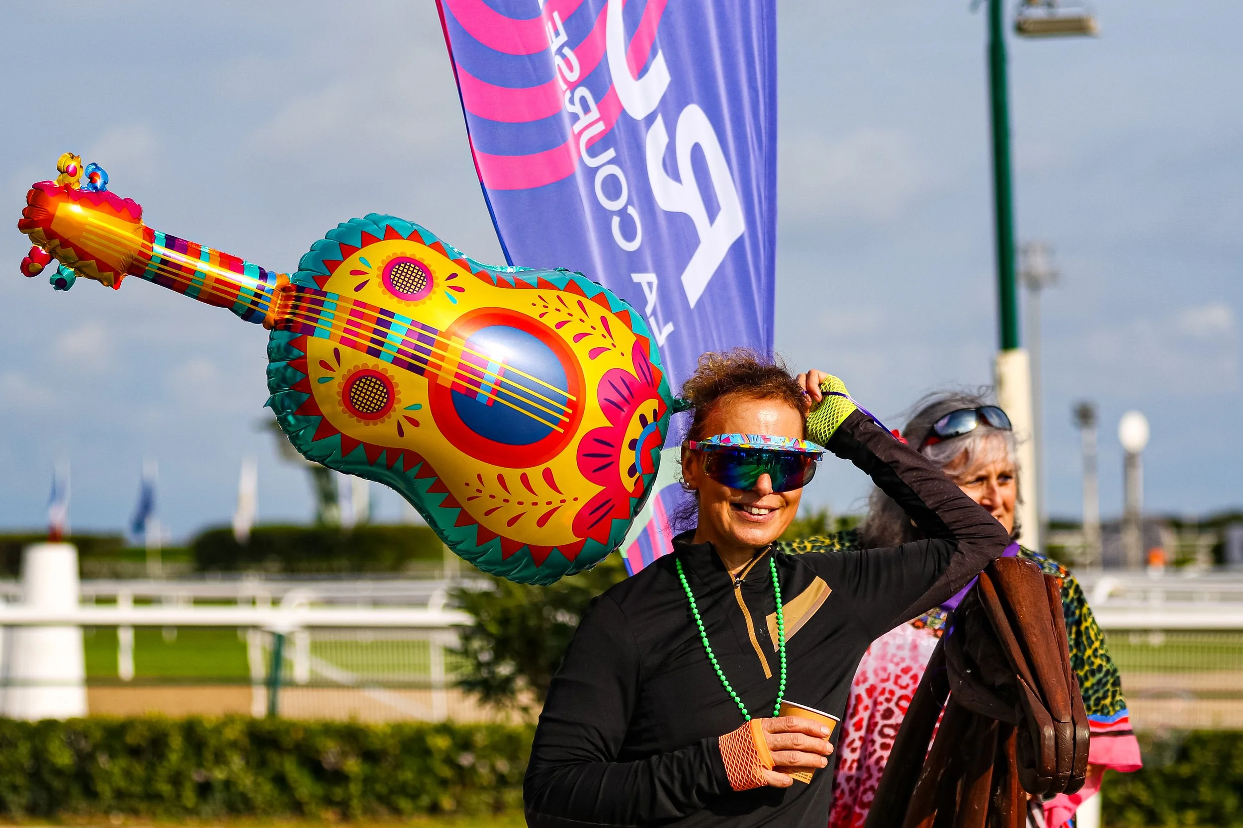Deux femmes souriantes portent des lunettes de soleil et des vêtements colorés, une d'elles tient un ballon en forme de guitare décoré, lors d'un événement en extérieur avec une bannière violette en arrière-plan.