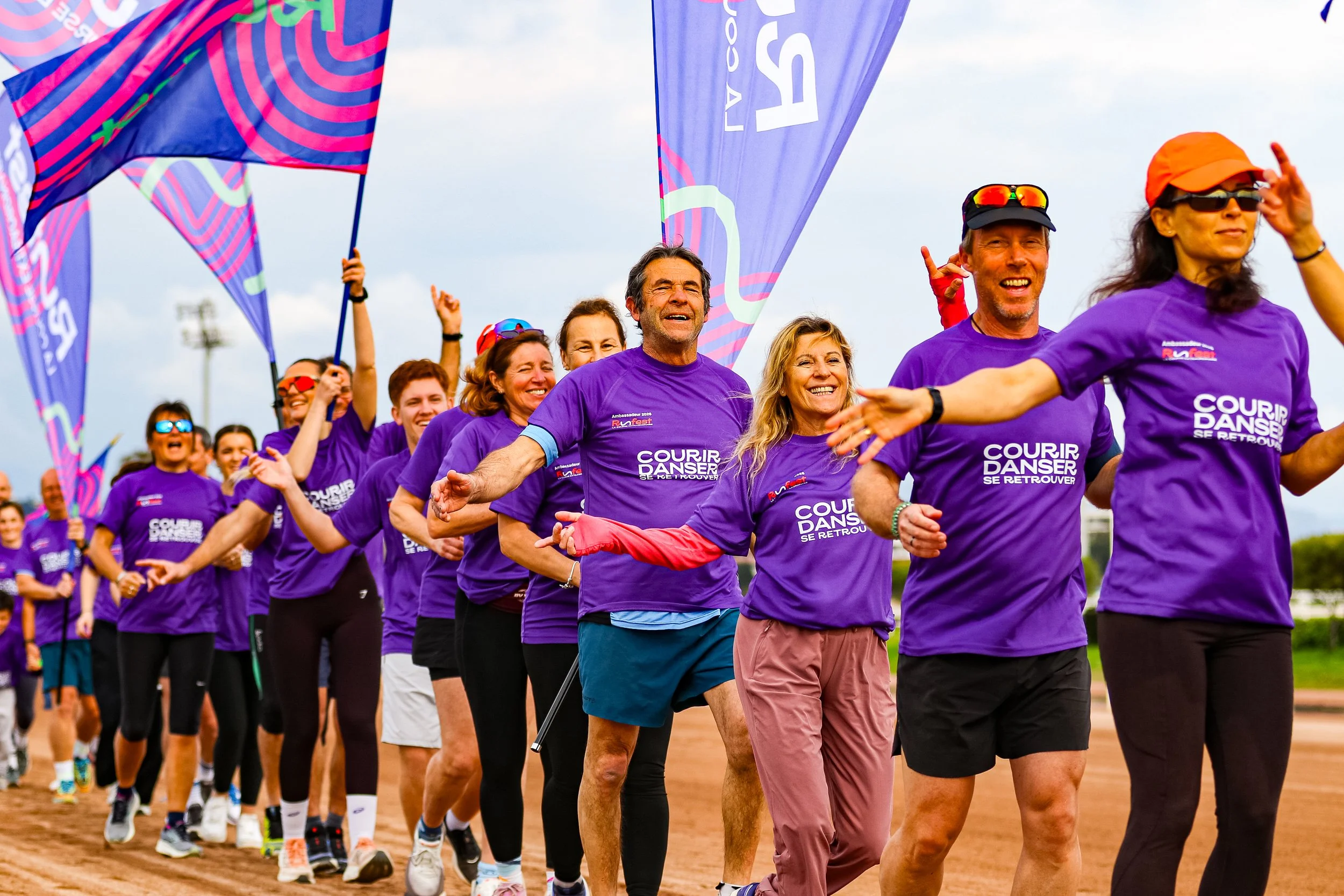 Groupe de personnes joyeuses participant à une course ou un événement sportif, portant des T-shirts violets avec le texte 'Courir, Danser, se retrouver', sur un terrain en plein air.