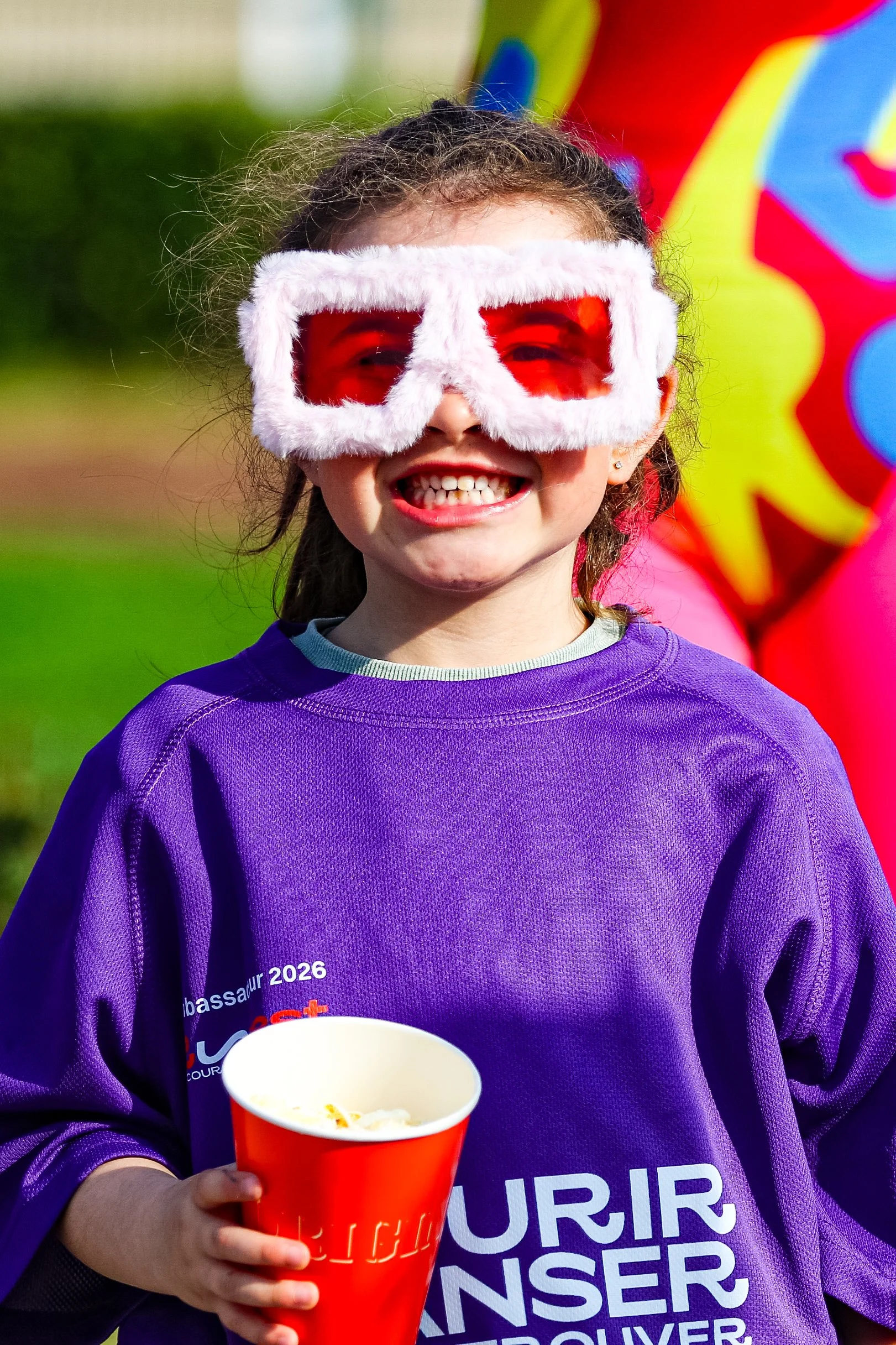 Jeune fille souriante portant des lunettes de soleil à moufles et un sweat violet, tenant un seau de popcorn, lors d'un événement en extérieur avec un ballon en arrière-plan.