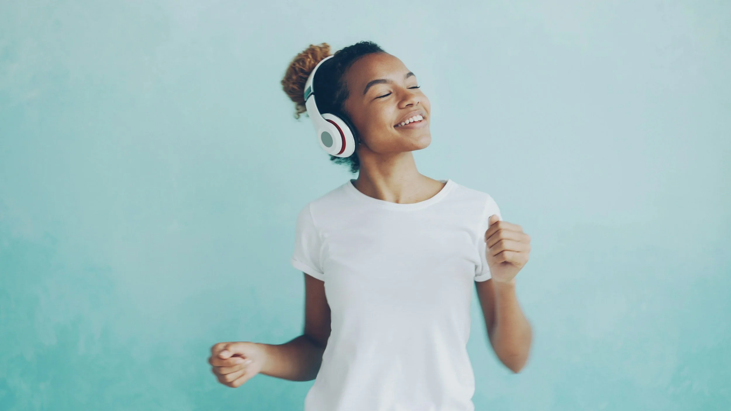 A happy young woman with curly hair wearing headphones and a white t-shirt, smiling and dancing against a light blue background.